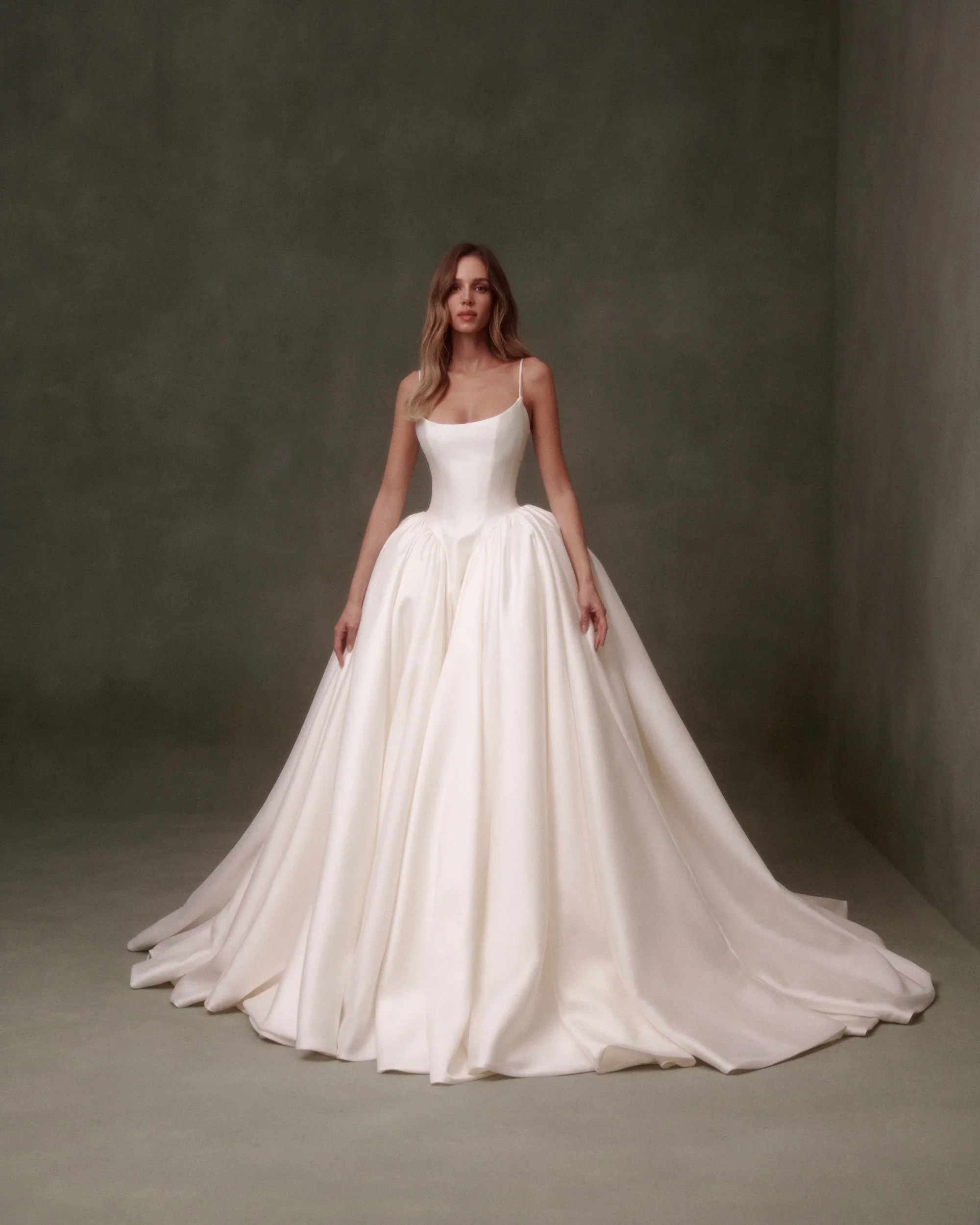 Model wearing a Nicole and Felicia minimalist satin ballgown wedding dress featuring delicate spaghetti straps, a square neckline, and a structured basque waist, standing against a dark olive backdrop.