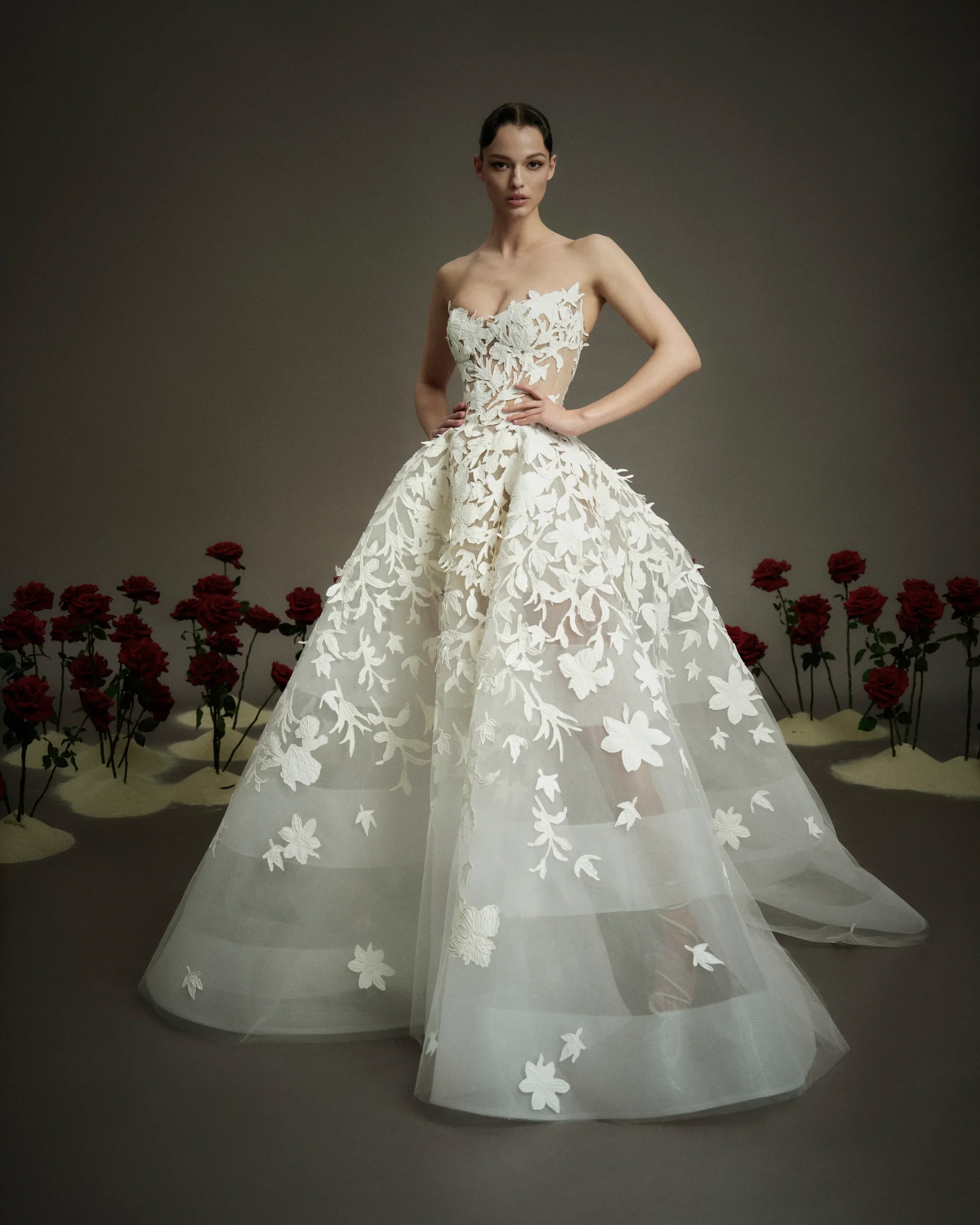 Model wearing a Nicole and Felicia strapless sheer wedding dress featuring intricate, oversized floral and vine appliqués cascading across a corset bodice and a full, banded tulle skirt, standing among red roses.