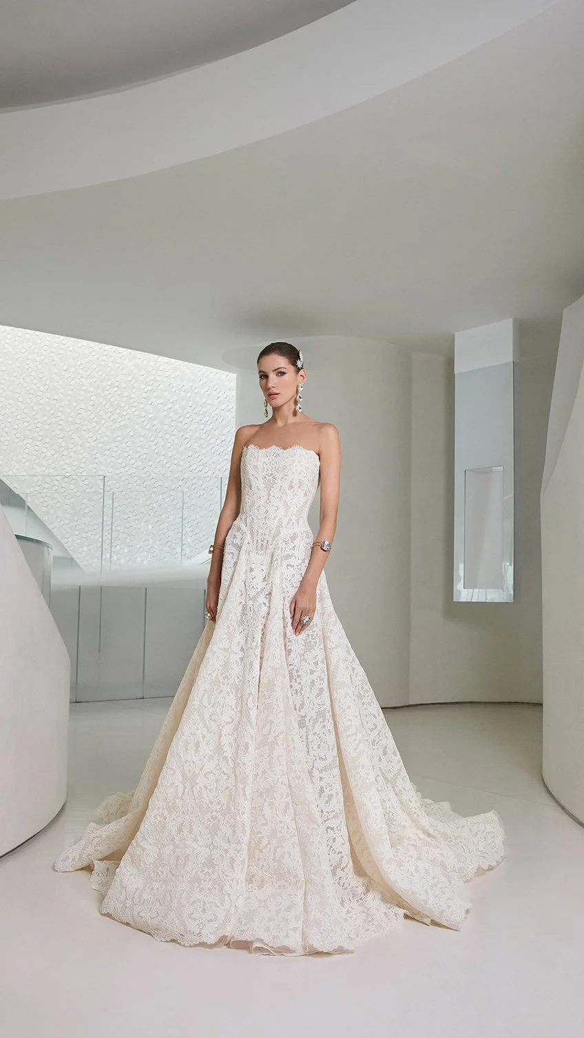 Full-length view of a model wearing a Galia Lahav strapless all-over lace wedding gown with a fitted waist and a flowing lace train, standing in a contemporary minimalist space.