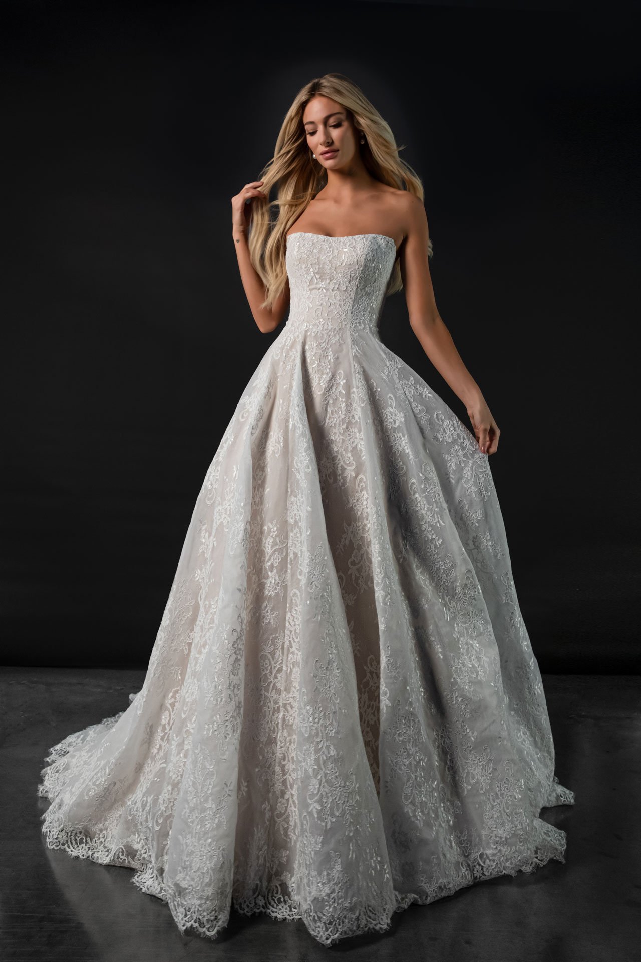 Model wearing a Martina Liana strapless lace fit-and-flare wedding gown, featuring a subtle peplum waist detail and a sweeping train, standing in a sunlit rustic stone hallway.