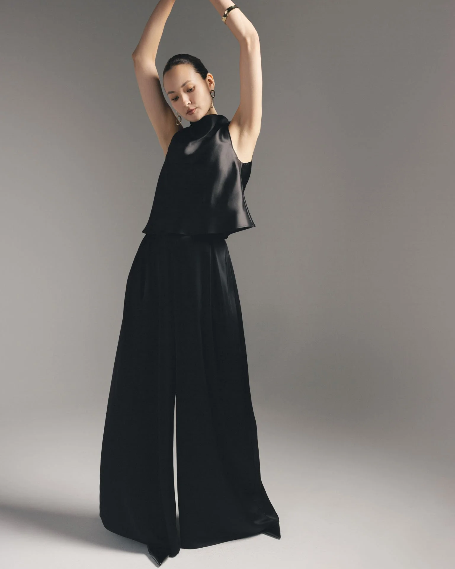 Model with arms raised wearing a Karis two-piece black silk set featuring a draped sleeveless top and high-waisted wide-leg trousers, styled with pointed black heels.