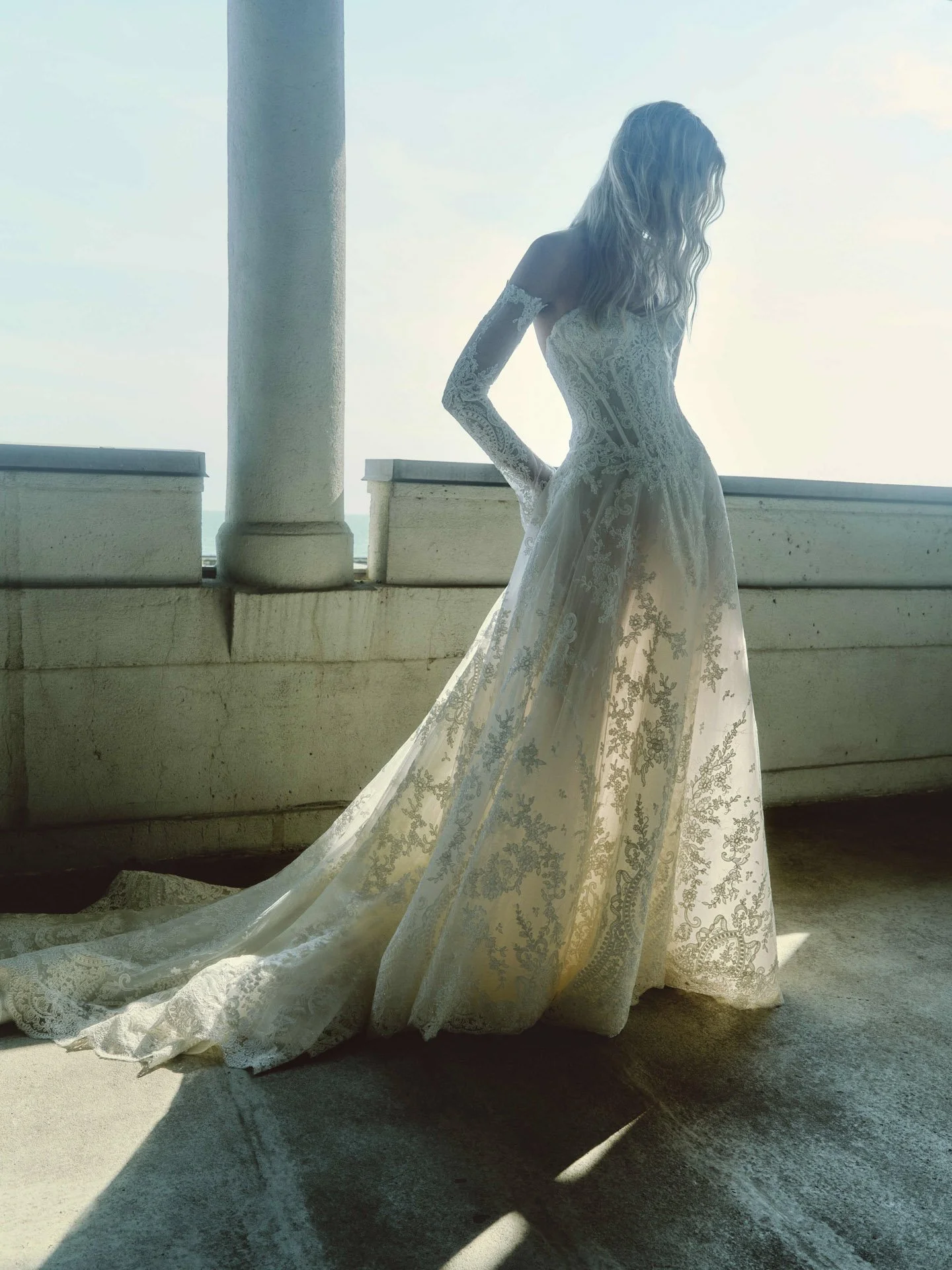 Model wearing a Rivini lace wedding dress featuring a structured basque waist corset bodice, an airy sheer lace skirt illuminated by sunlight, and matching detached off-the-shoulder long lace sleeves.