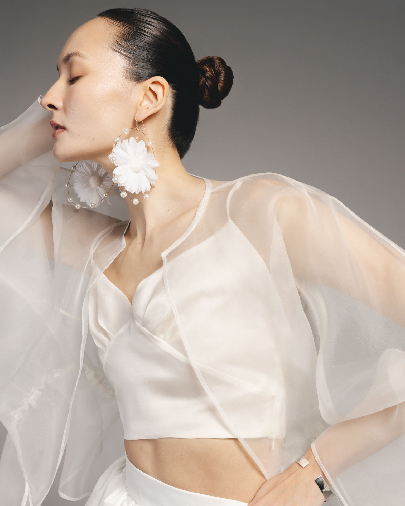 Close-up of a model wearing a Karis white silk sweetheart crop top layered under a sheer white organza jacket, styled with oversized white floral hoop earrings.