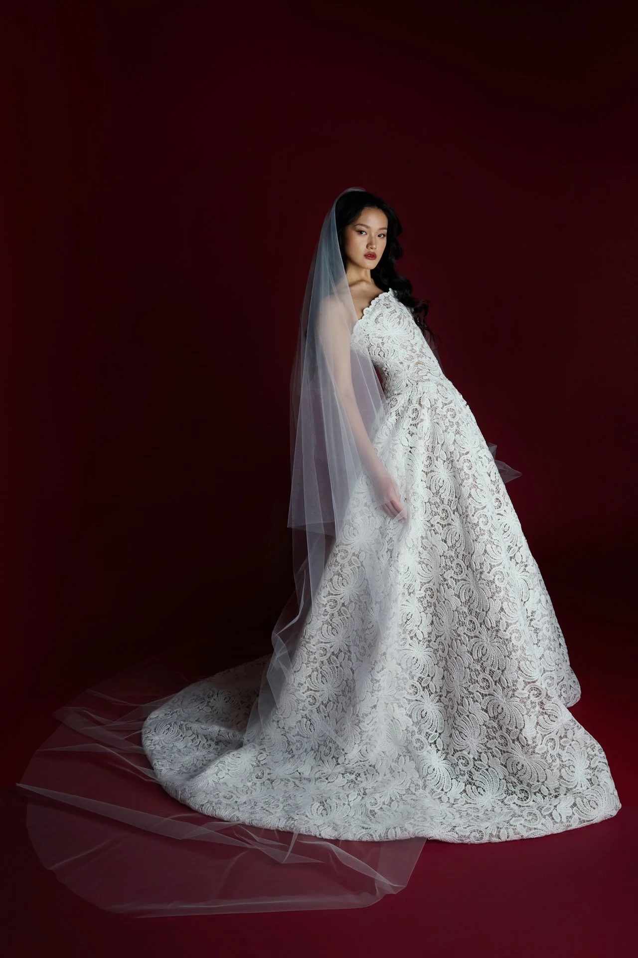 Full-length view of a model wearing a Jaclyn Whyte strapless lace wedding dress with a voluminous A-line skirt and a long lace train, styled with a sheer, flowing tulle veil against a dark red backdrop.