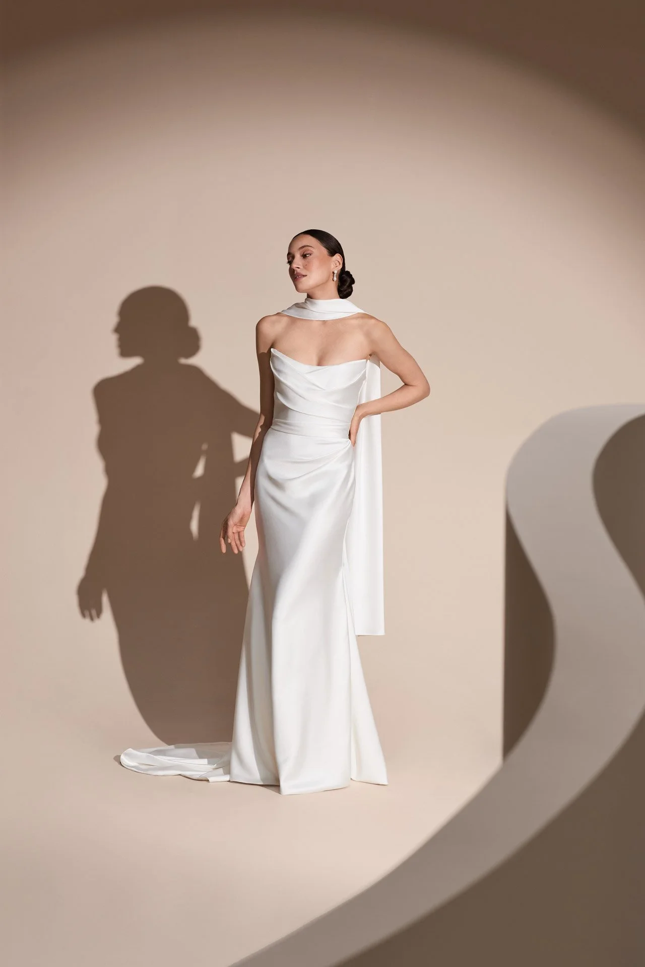 Model wearing a Savannah Miller strapless white satin sheath wedding dress with a draped cowl neckline, fitted waist, and an attached neck scarf trailing down the back against a warm studio backdrop.