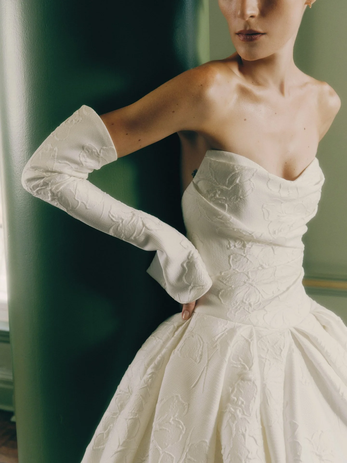 Close-up of a model wearing a Rivini strapless ballgown wedding dress crafted from textured floral embossed fabric, featuring an elegantly draped bodice and matching full-length opera gloves.