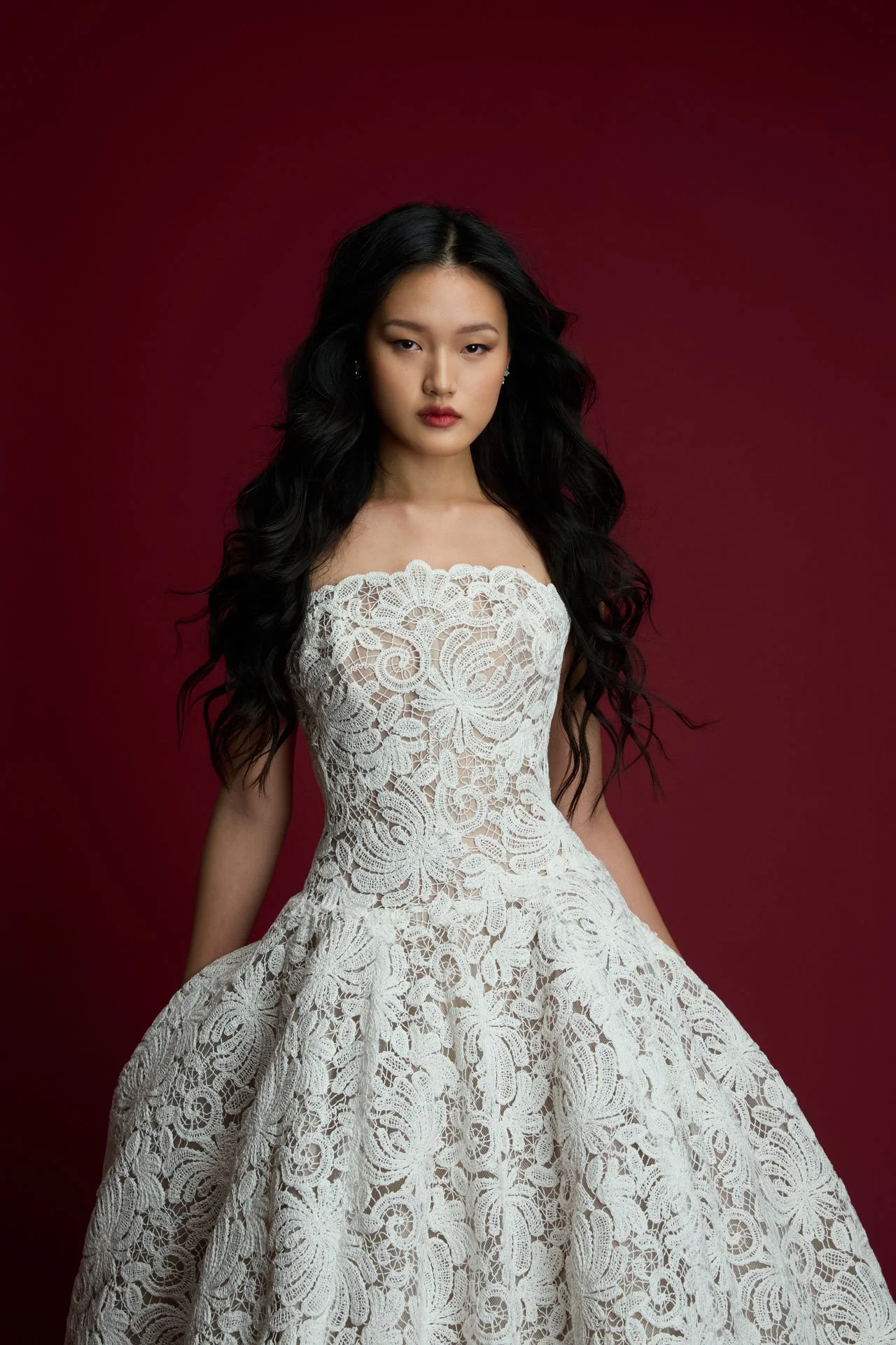 Model wearing a Jaclyn Whyte strapless lace ballgown wedding dress featuring a fitted bodice with bold, textured all-over botanical lace, standing against a deep crimson red background.