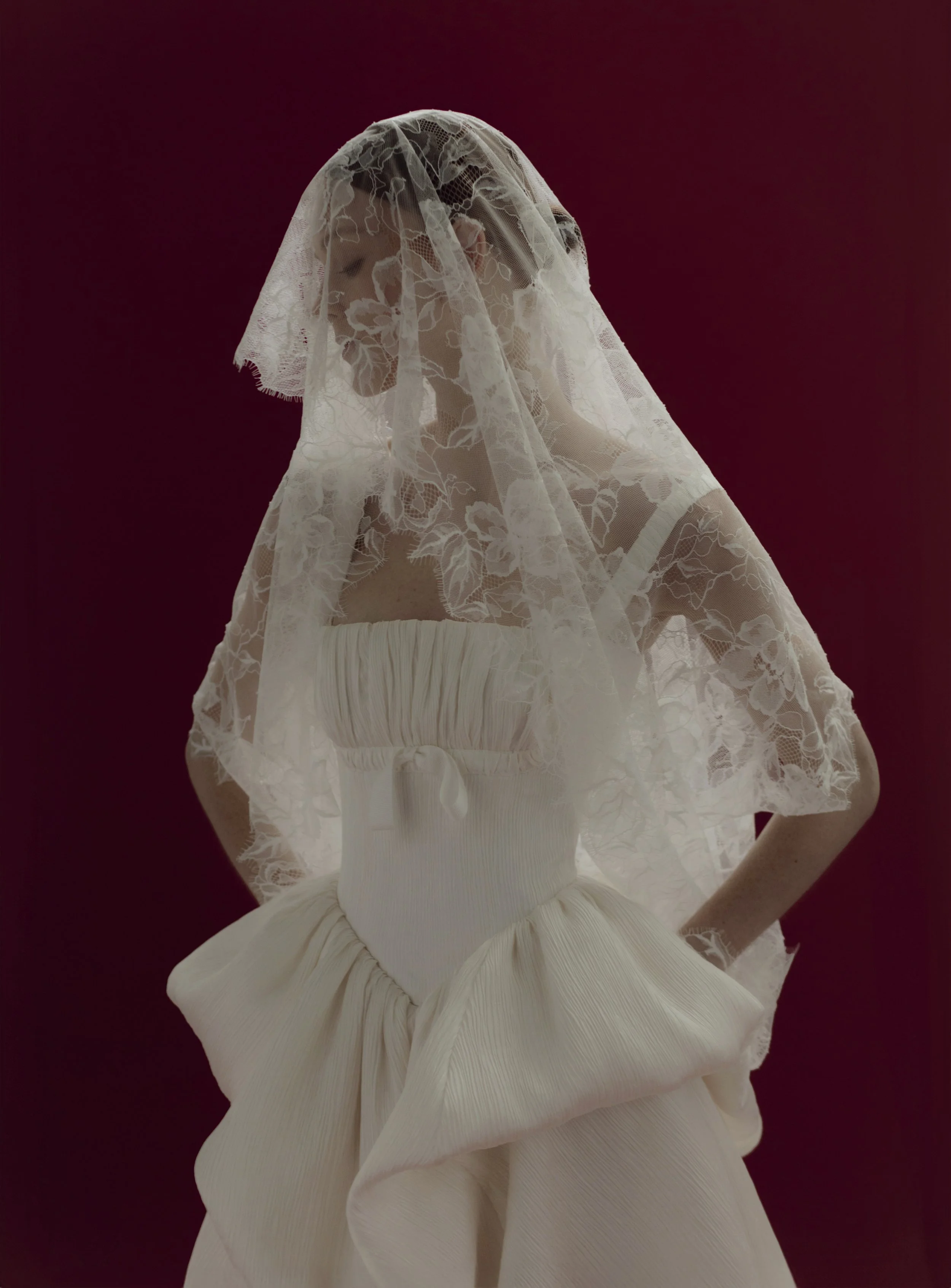 Model wearing a whimsical OUMA strapless drop-waist ballgown featuring a vertically pleated basque corset bodice, styled with a delicate floral lace veil covering her face against a dark red background.