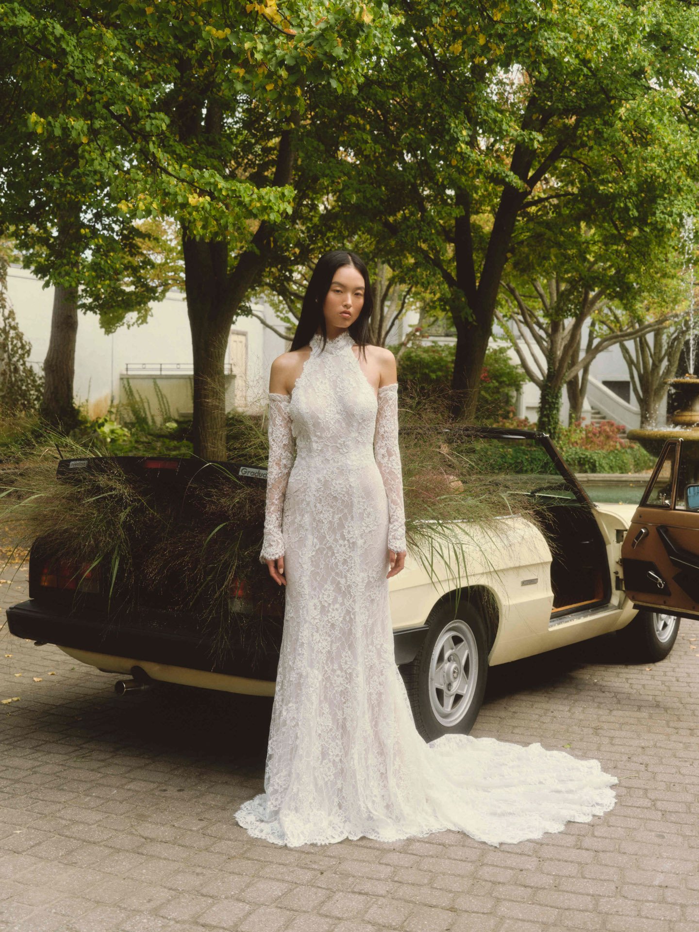 Model wearing an Alyne all-over lace halter sheath wedding dress with long fitted sleeves and a sweep train, standing outdoors next to a vintage beige convertible car filled with dried grass and foliage.