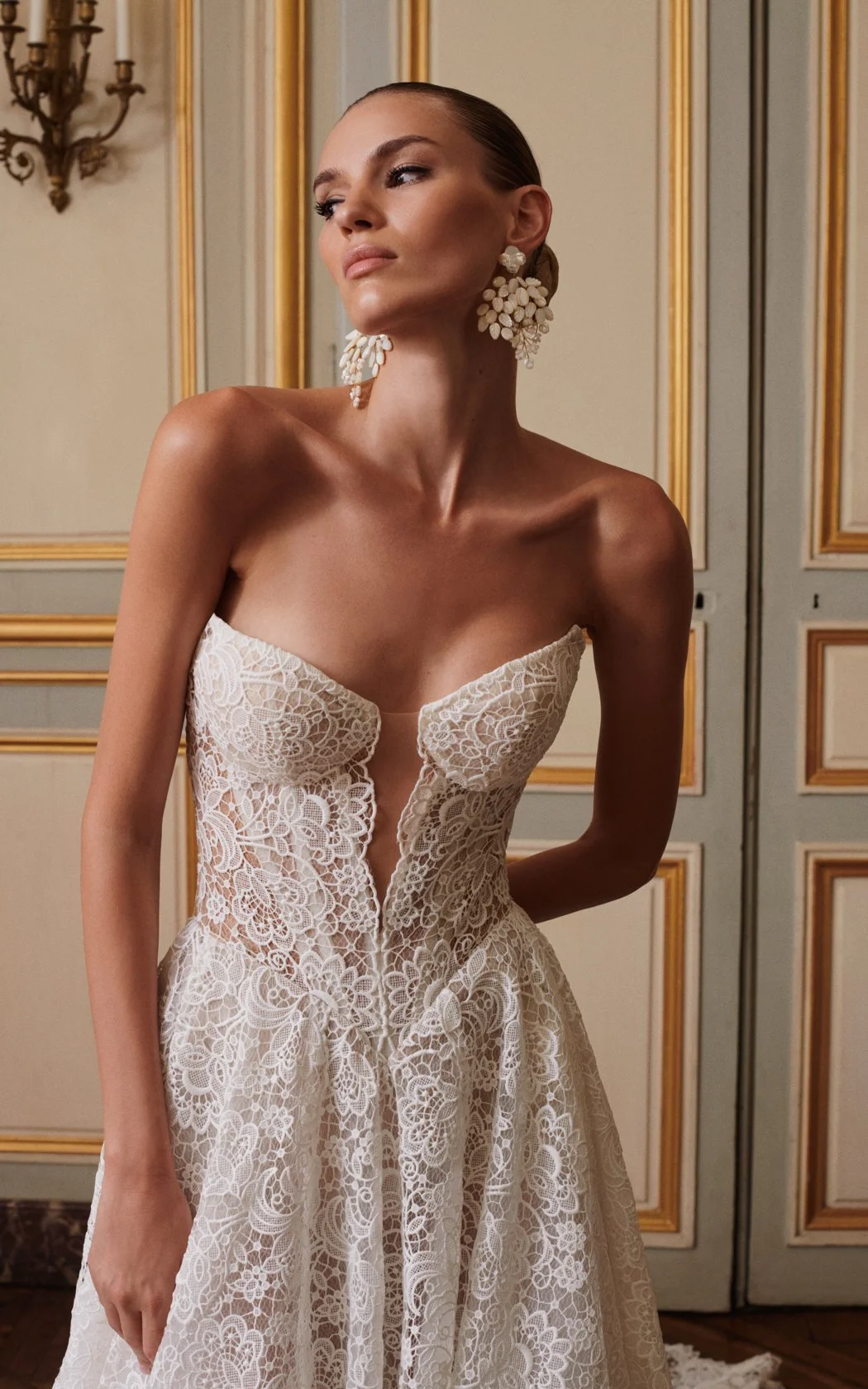 Close-up of a model wearing a Galia Lahav strapless lace wedding dress, highlighting a structured corset bodice with a deep V illusion plunge neckline and large floral statement earrings.