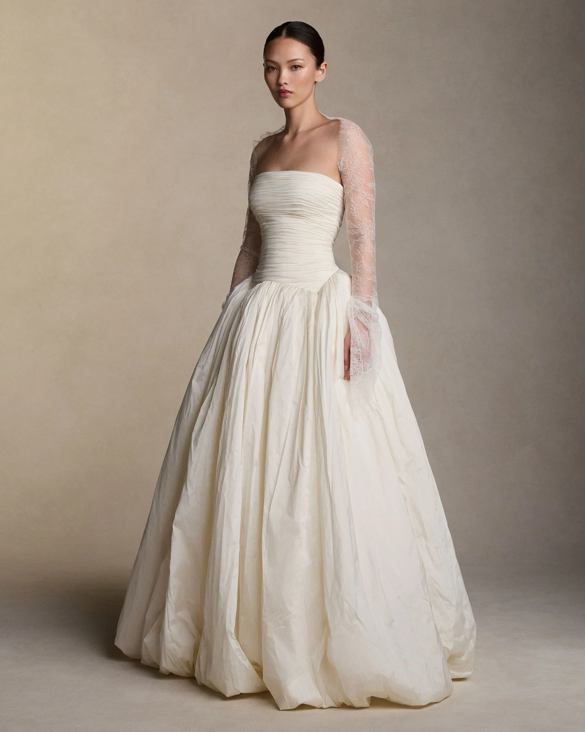 Model wearing a Danielle Frankel drop-waist ballgown wedding dress showcasing a ruched strapless bodice layered with sheer long lace sleeves and a full textured skirt.