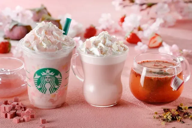 Three beverages on a pink surface with whipped cream topping, two in Starbucks cups and one in a clear glass cup, with strawberries and pink flowers in the background.