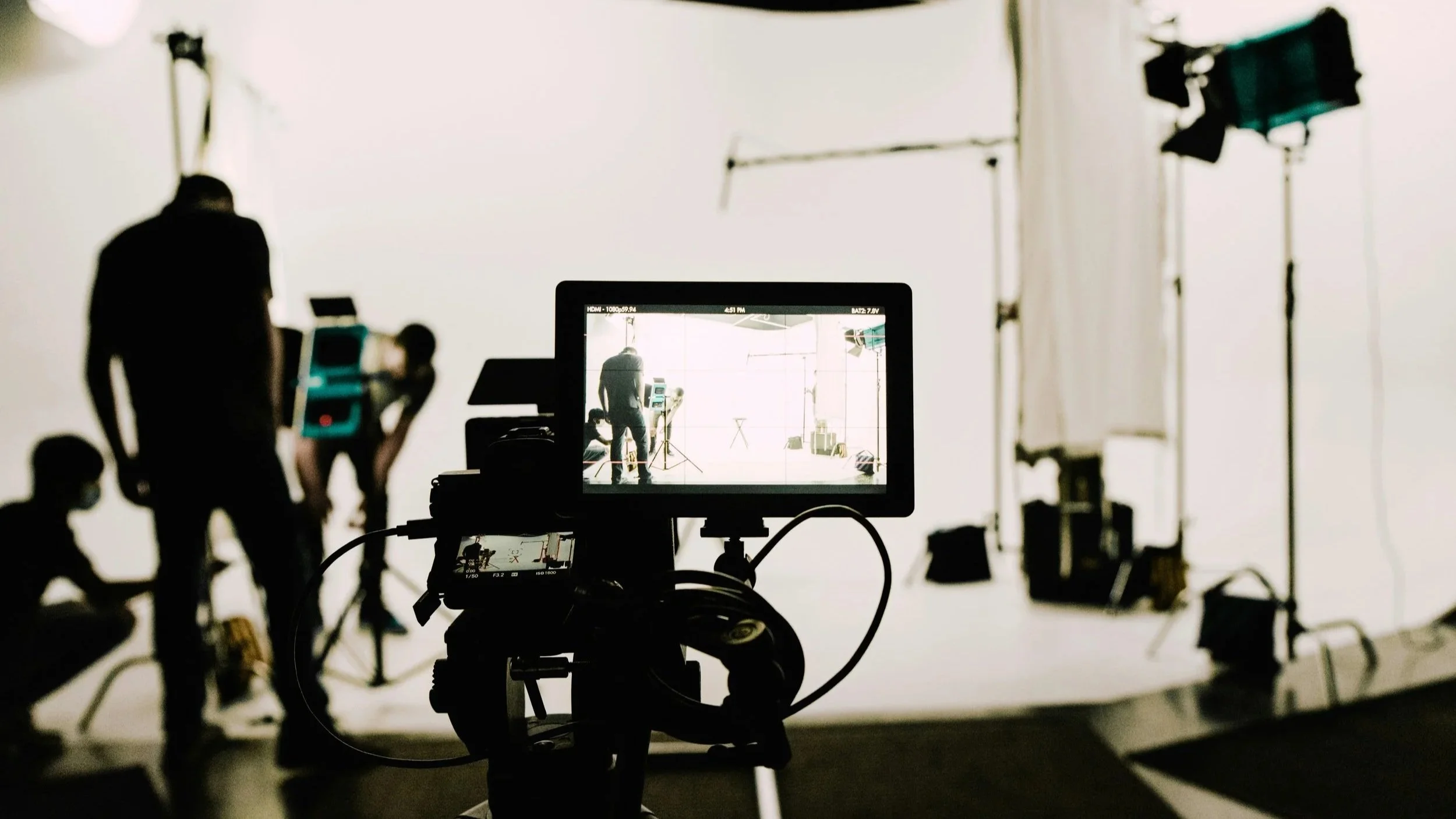 A Producer's perspective, behind-the-scenes view, of a video shoot with a camera on a tripod, monitor displaying the scene, and crew members adjusting the set-up in a white studio space with lighting equipment and a white backdrop.