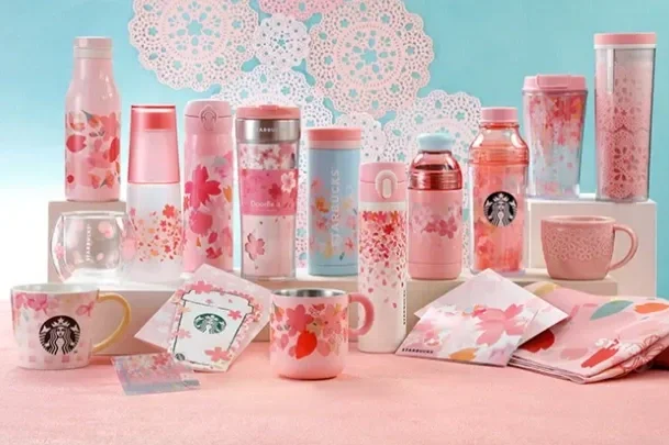 A collection of pink and floral Starbucks tumblers, mugs, and napkins displayed on a table with a blue and white floral background.
