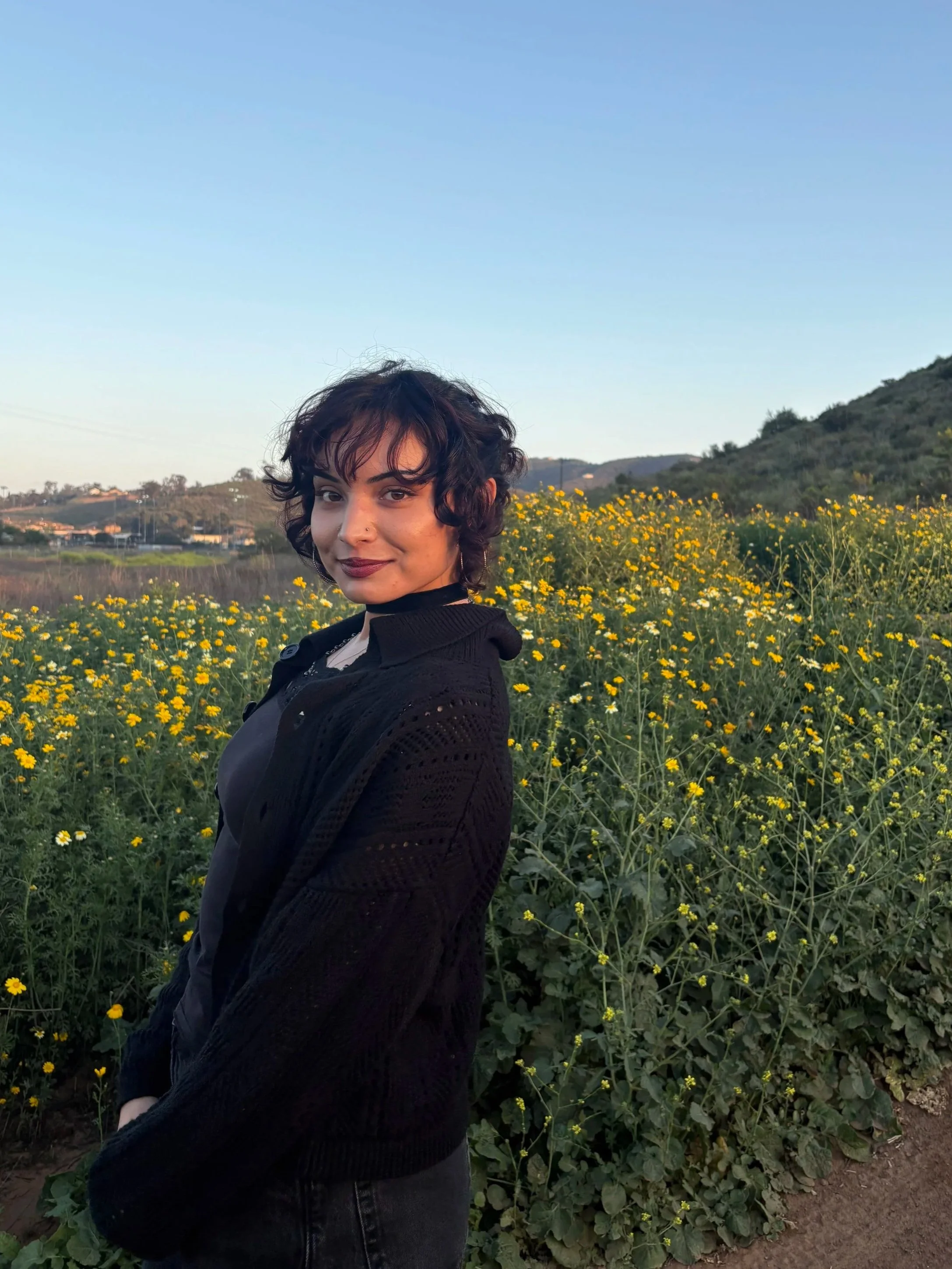 A woman with short, curly dark hair and light makeup standing in a field of yellow flowers with hills in the background during the daytime.