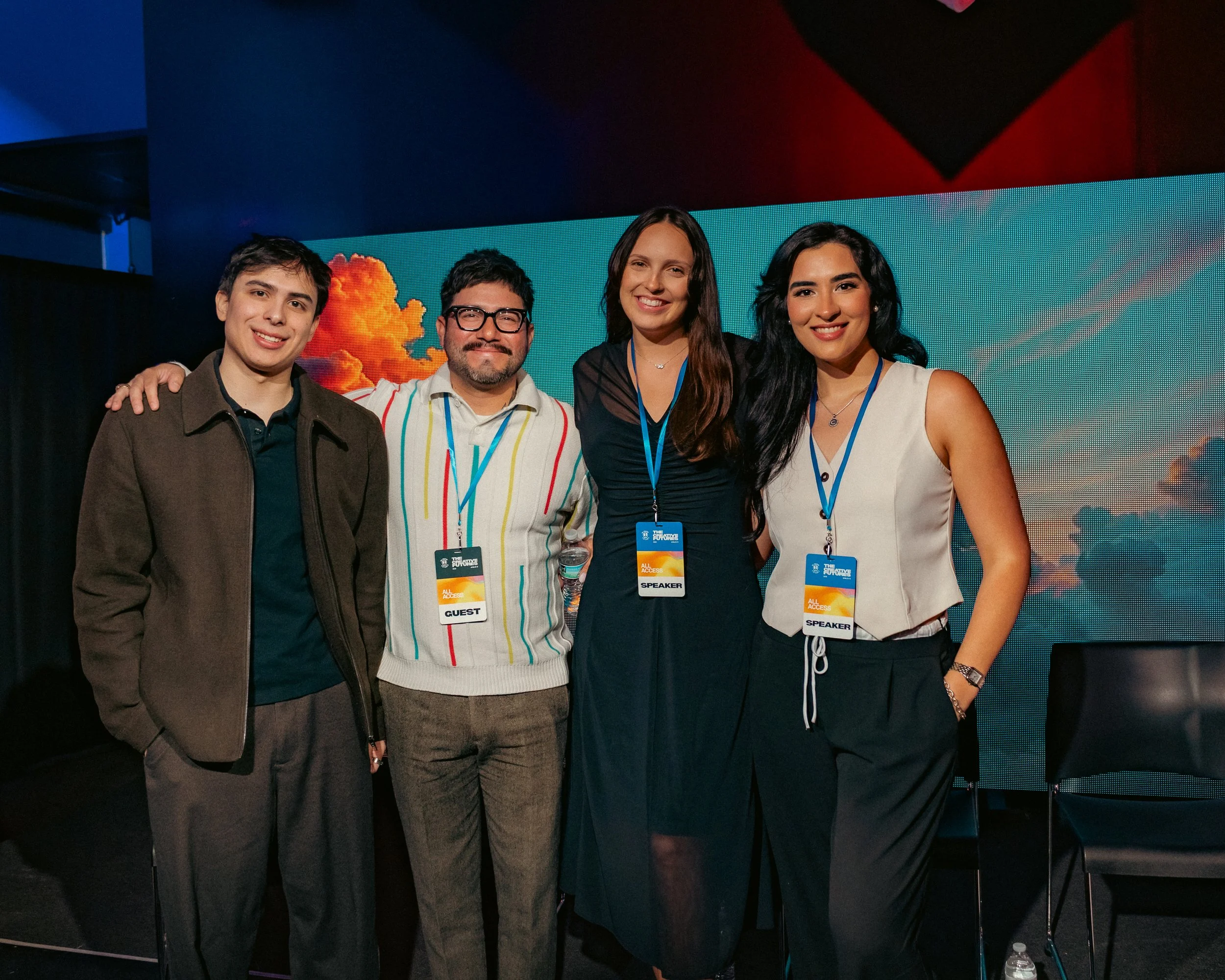 Four people standing together at an event, wearing badges. One man and three women, smiling in front of a large digital screen displaying a background with red and black geometric shapes and a cloud of orange smoke. The men and women are dressed in casual and semi-formal attire.