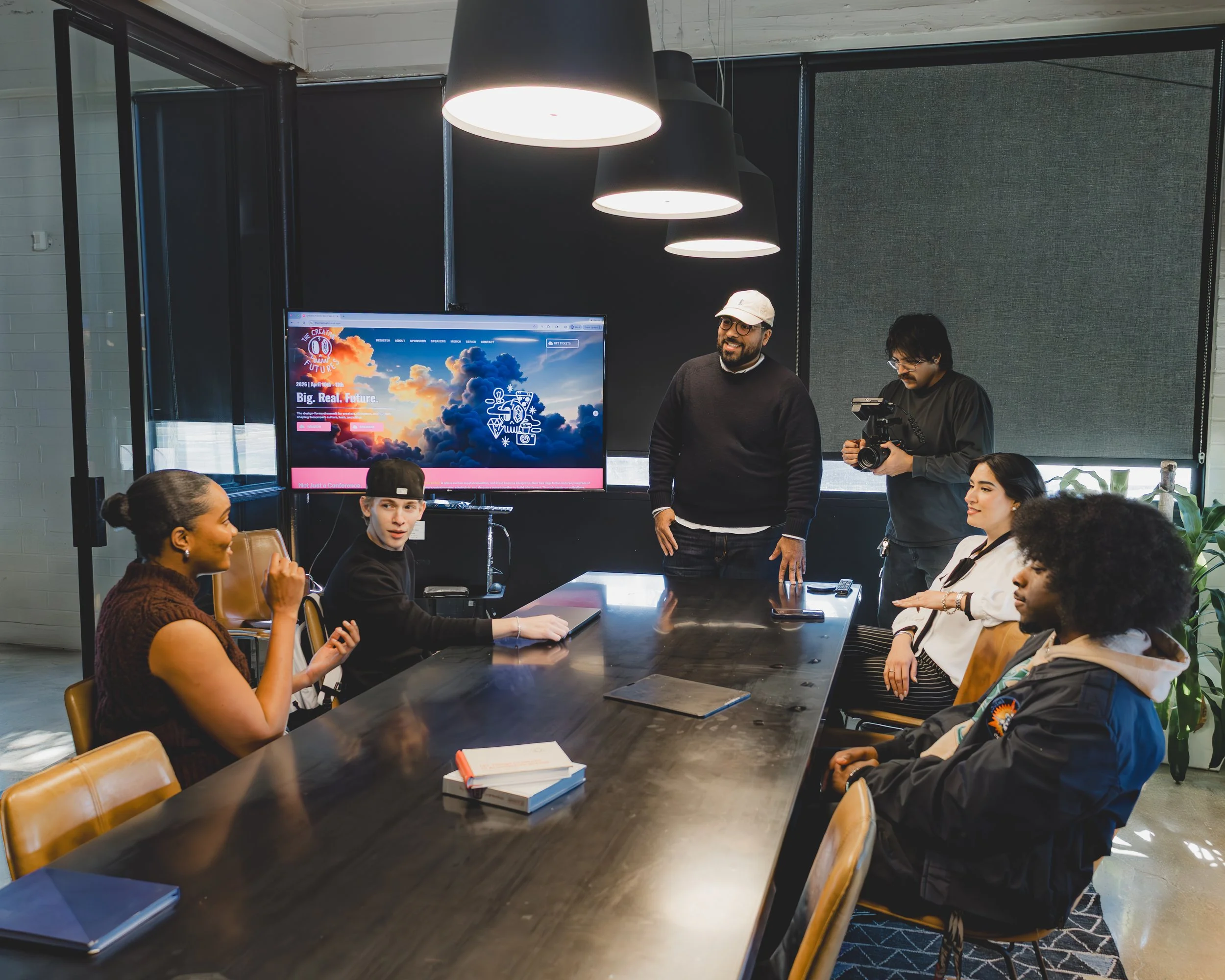 A diverse group of people sits around a conference table in a modern office, engaging in a meeting. Some participants are listening attentively, while one woman gestures as she speaks. There is a large screen at the front displaying a colorful cloud-themed presentation. Two men stand near the screen, one smiling and the other holding a camera. The room is well-lit with ceiling lamps, and there are laptops, notebooks, and books on the table. Large windows with blinds are in the background.