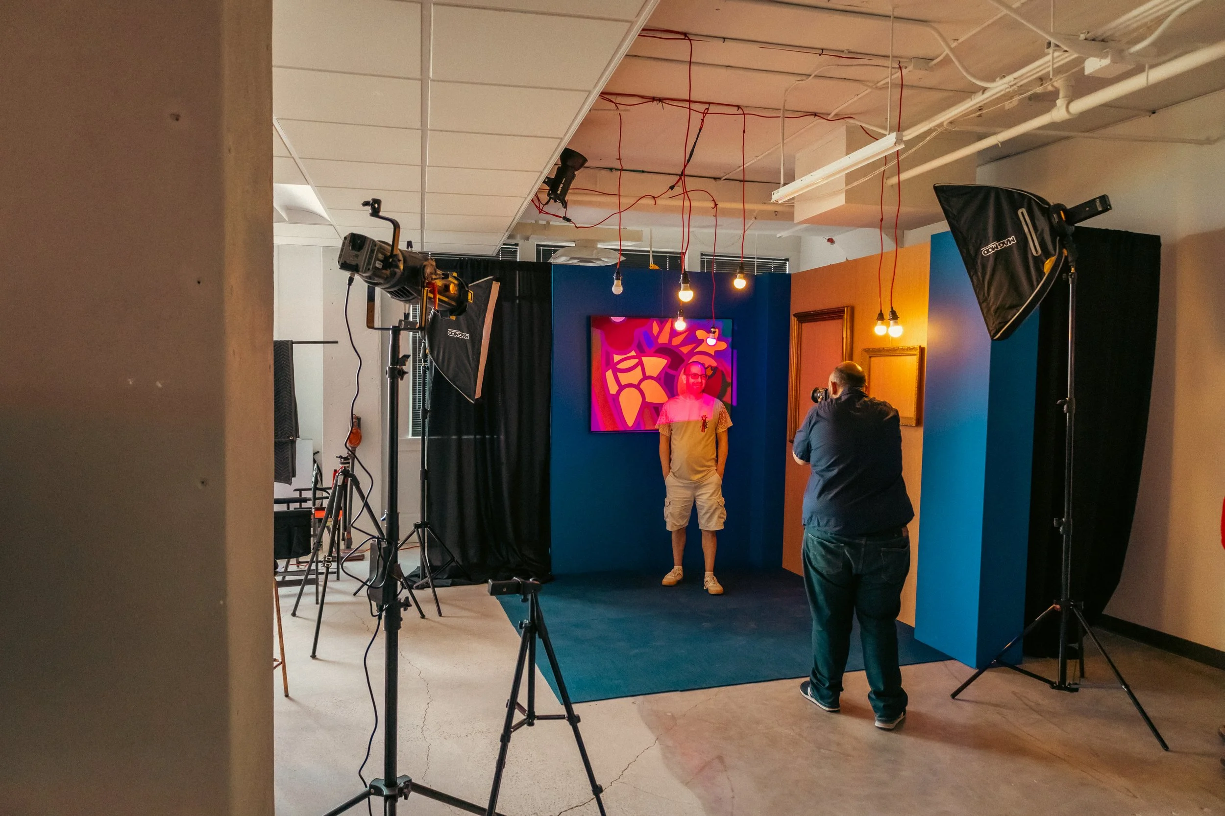 A photography studio setup with lighting equipment, a blue backdrop, and a colorful abstract digital artwork on the wall. Three people are present, one standing in front of the artwork, another photographing, and the third adjusting the lights. Exposed ceiling pipes and red electrical wires are visible.