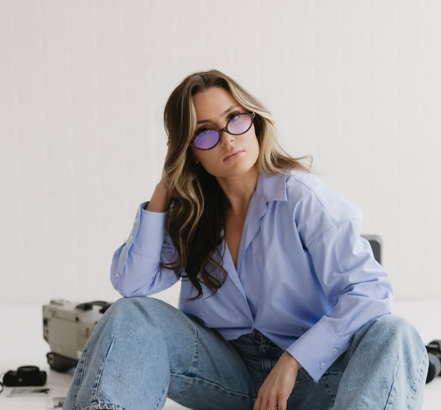 A young woman with wavy hair, wearing purple sunglasses and a light blue button-up shirt, sitting with her legs crossed and looking at the camera.