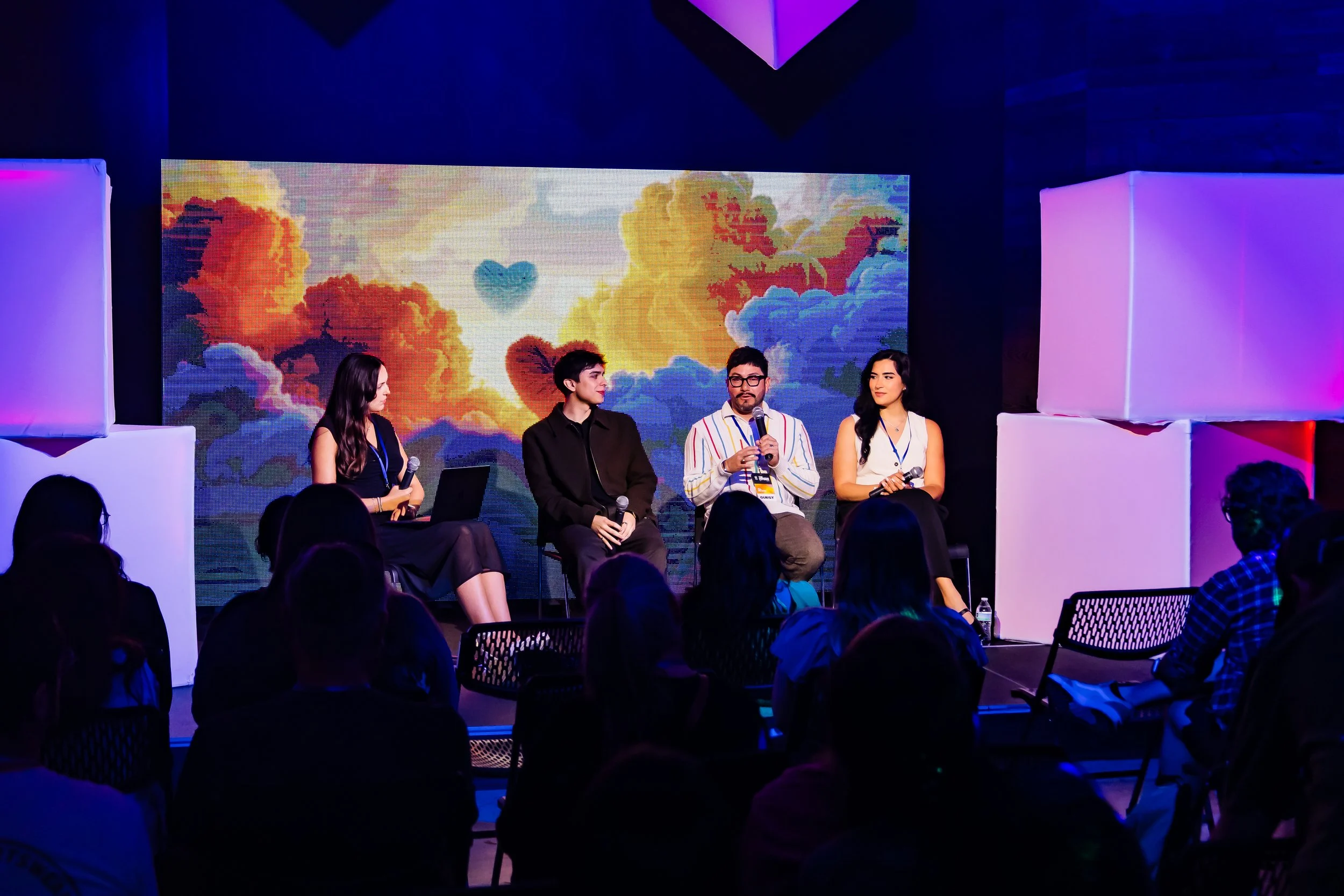 Four individuals sitting on a stage in front of a colorful digital backdrop featuring hearts and clouds, speaking into microphones at a conference or panel event, with an audience seated in front.