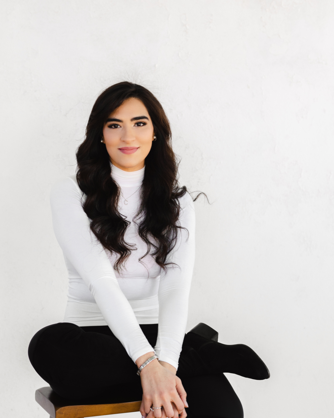 A woman with long dark wavy hair, wearing a white turtleneck and black pants, seated on a wooden stool against a white textured wall.