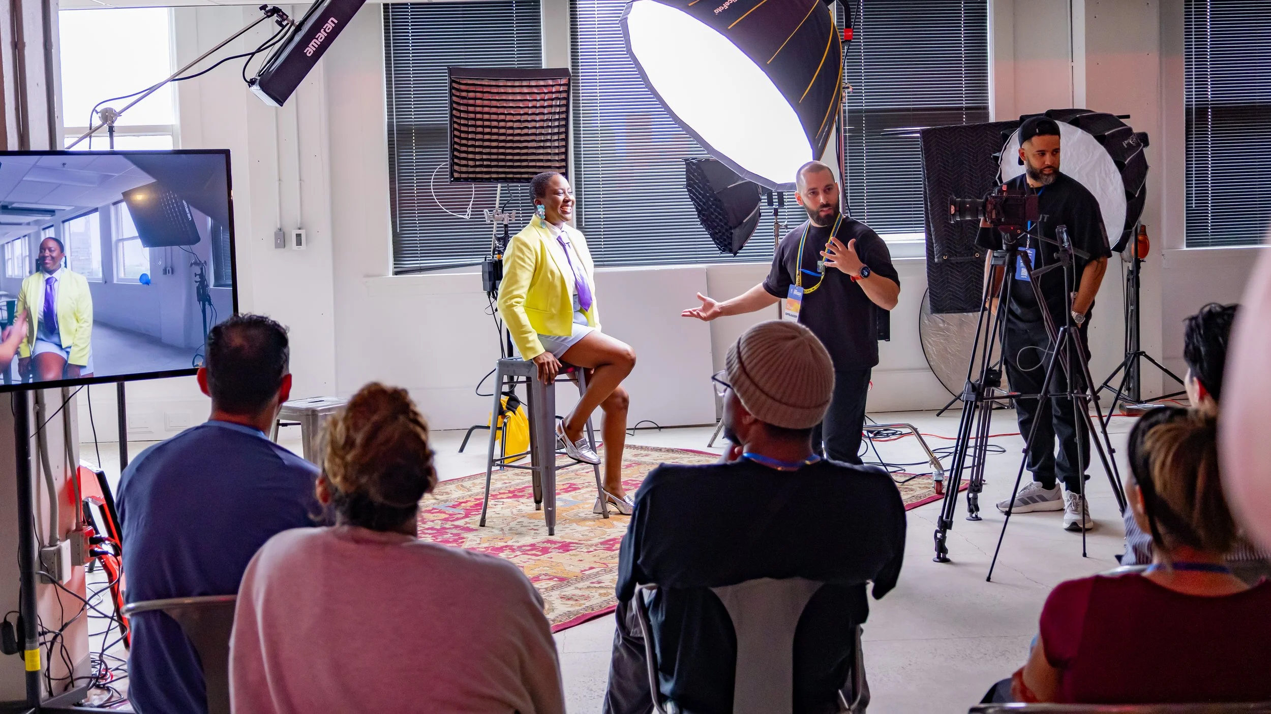 A woman in a bright yellow blazer sitting on a stool during a recording or interview, with a man standing next to her explaining something. There are professional lighting and camera equipment set up, and an audience watching.