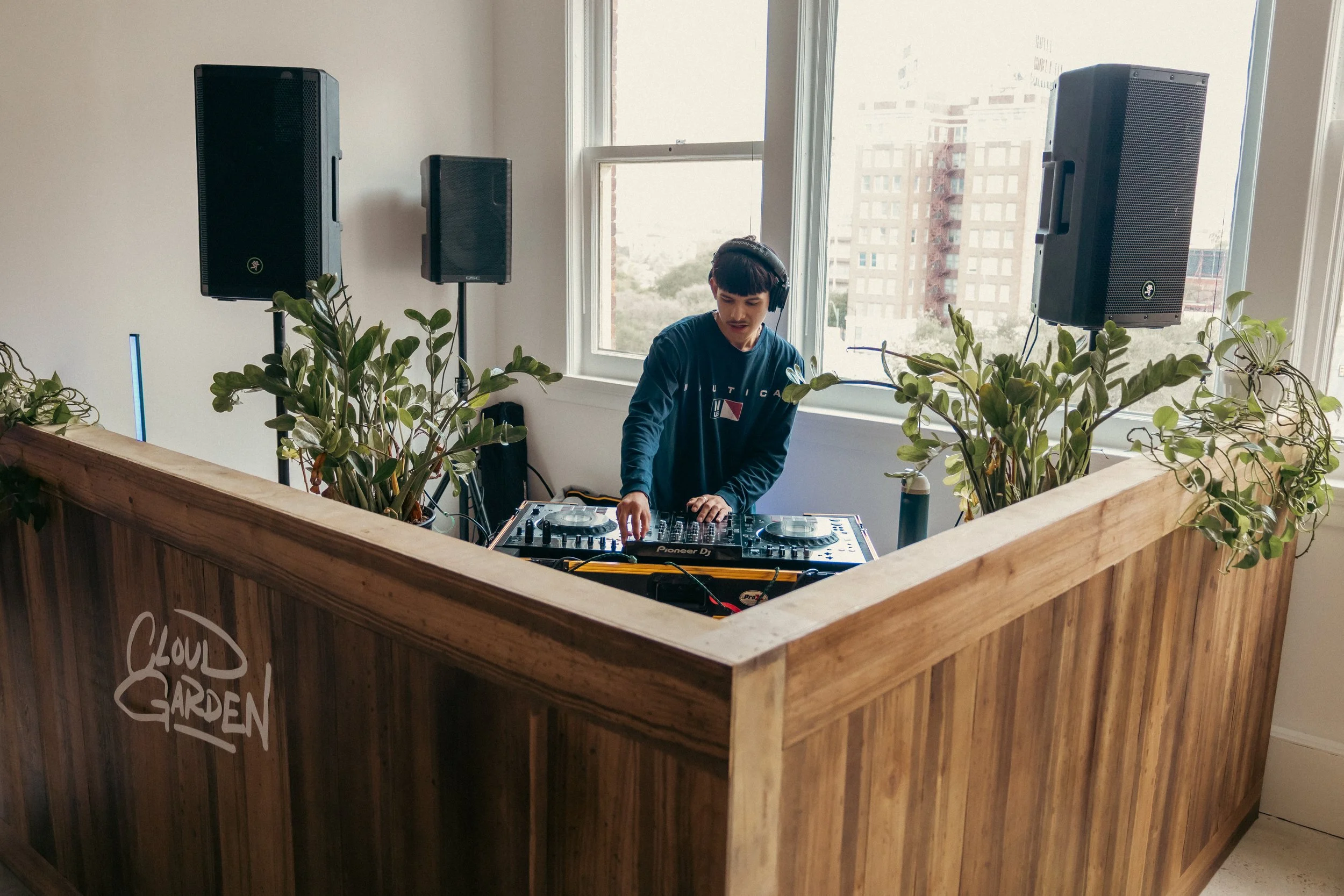 A young DJ wearing headphones is set up with a turntable and mixer inside a wooden booth with greenery on the sides, overlooking a cityscape through large windows.