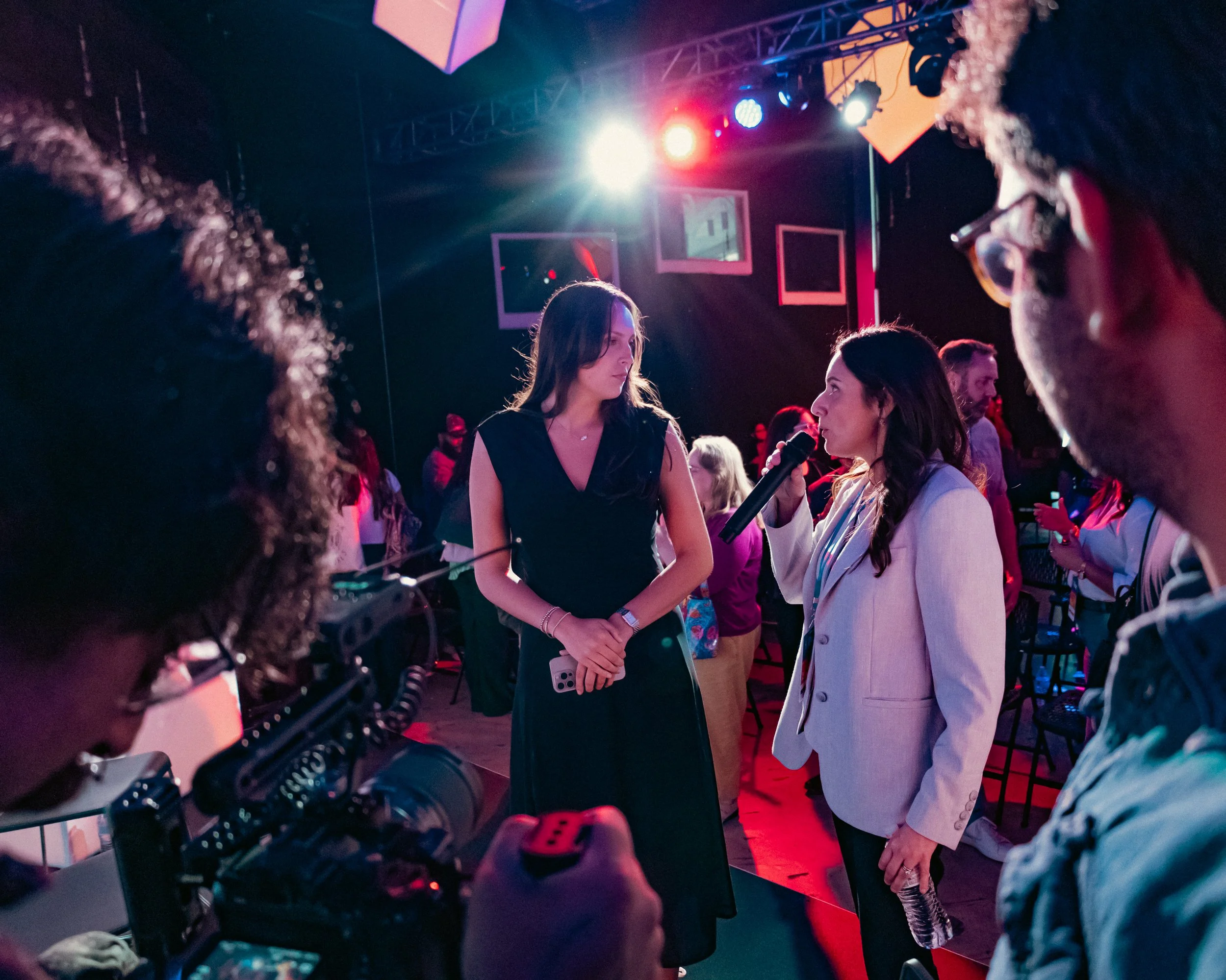 A group of women engaged in conversation at an indoor event with colorful stage lighting, including a woman with a microphone and a woman with a headset, amid multiple hook-shaped light fixtures.