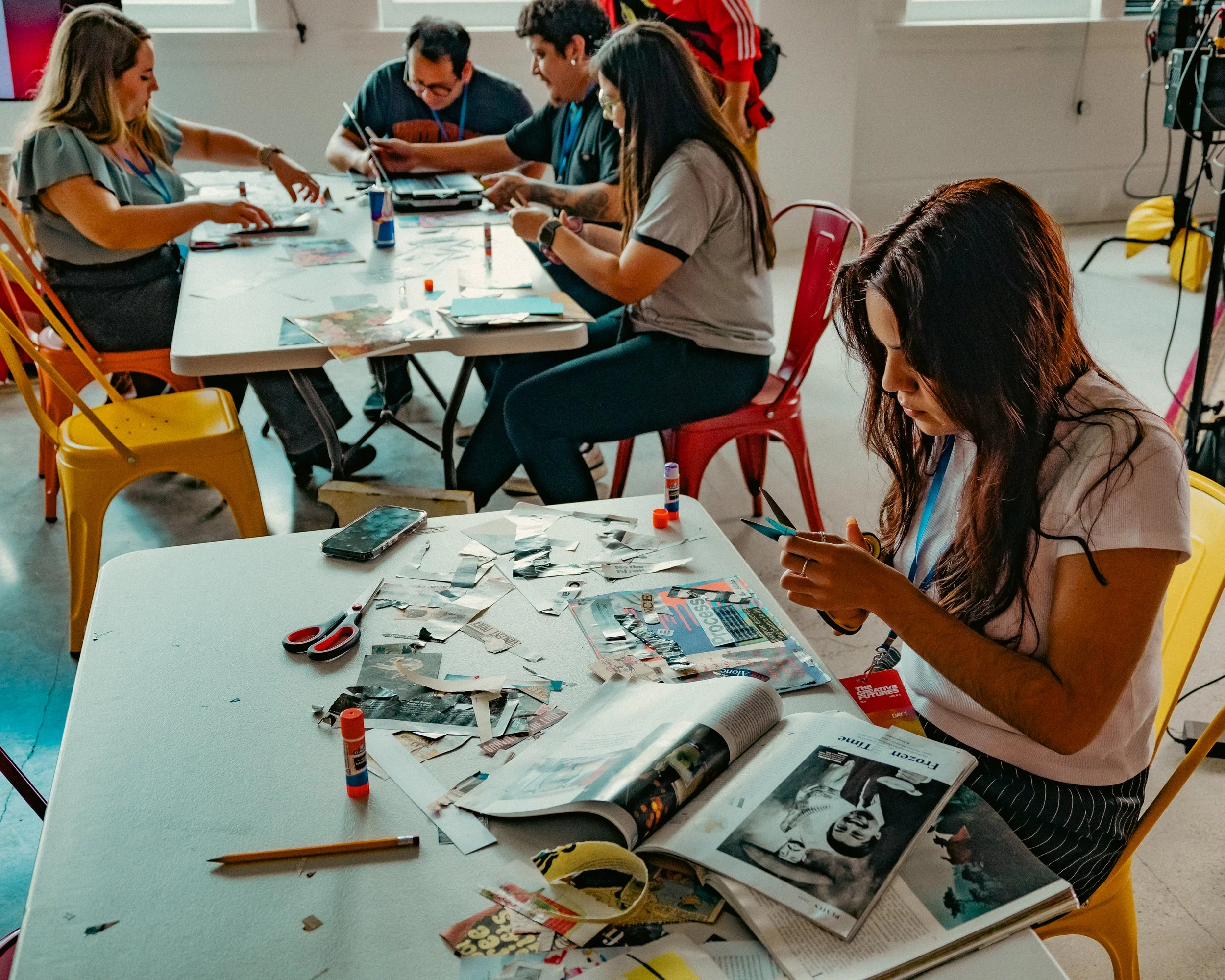 A group of five people working on a collage or craft project at a long white table in a bright room. They are surrounded by scissors, glue sticks, magazines, and newspapers. The people are engaged and focused on their work, with some sitting and one standing. The table is cluttered with craft supplies and paper cutouts.