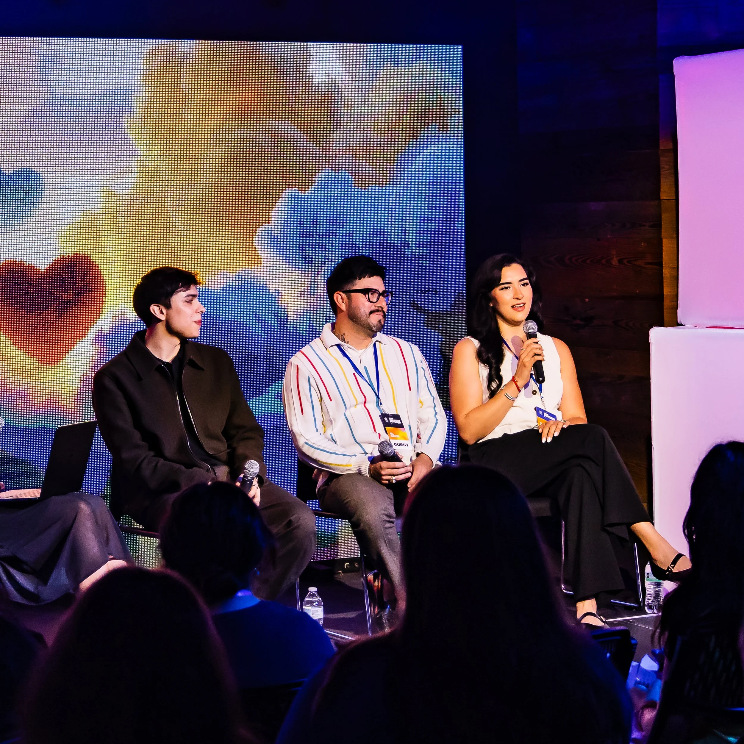 People sitting on a stage during a panel discussion at an event, with a large colorful digital screen in the background showing clouds and a heart symbol.