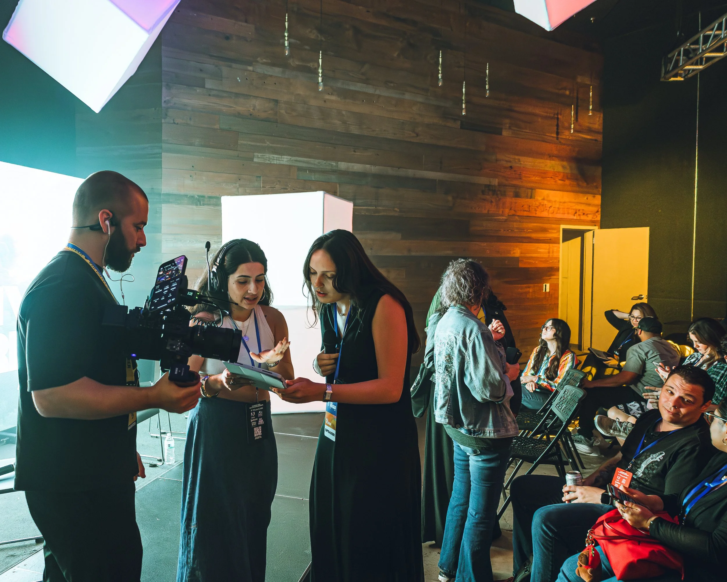 People gathered at an event, some seated and some standing, with a wooden wall background. Two women in the center appear to be discussing or working on a project on a tablet, assisted by a man with a camera or filming equipment. Other attendees are engaged with their devices or in conversation.