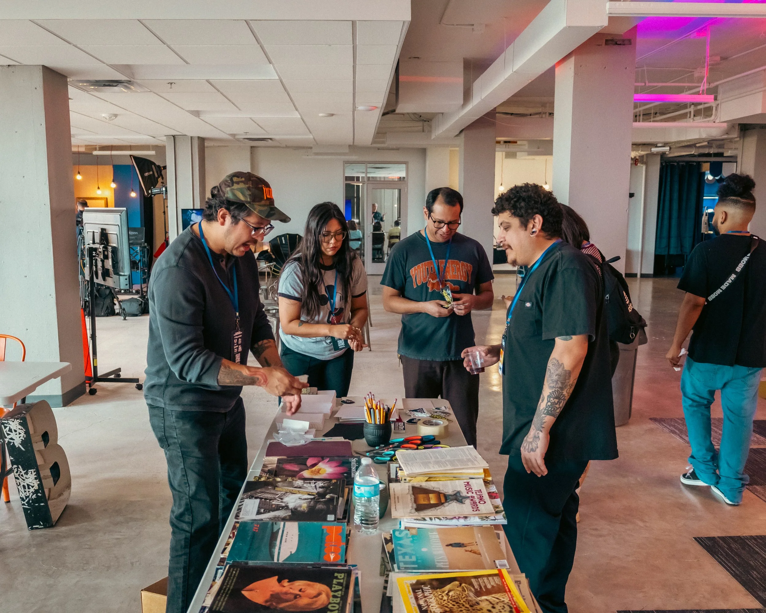 A group of five people gathered around a table with books, magazines, pens, and a water bottle at an indoor event. They appear to be engaging in a discussion or activity.