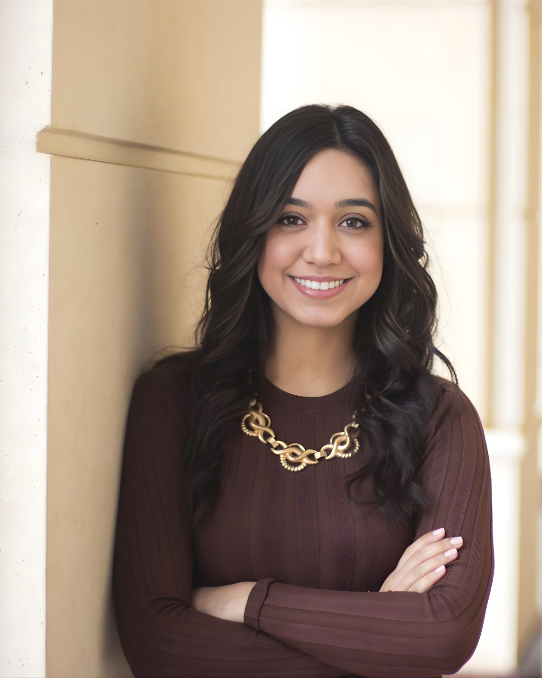 A young woman with long dark wavy hair, smiling, standing beside a wall with her arms crossed, dressed in a brown top and a gold chain necklace.