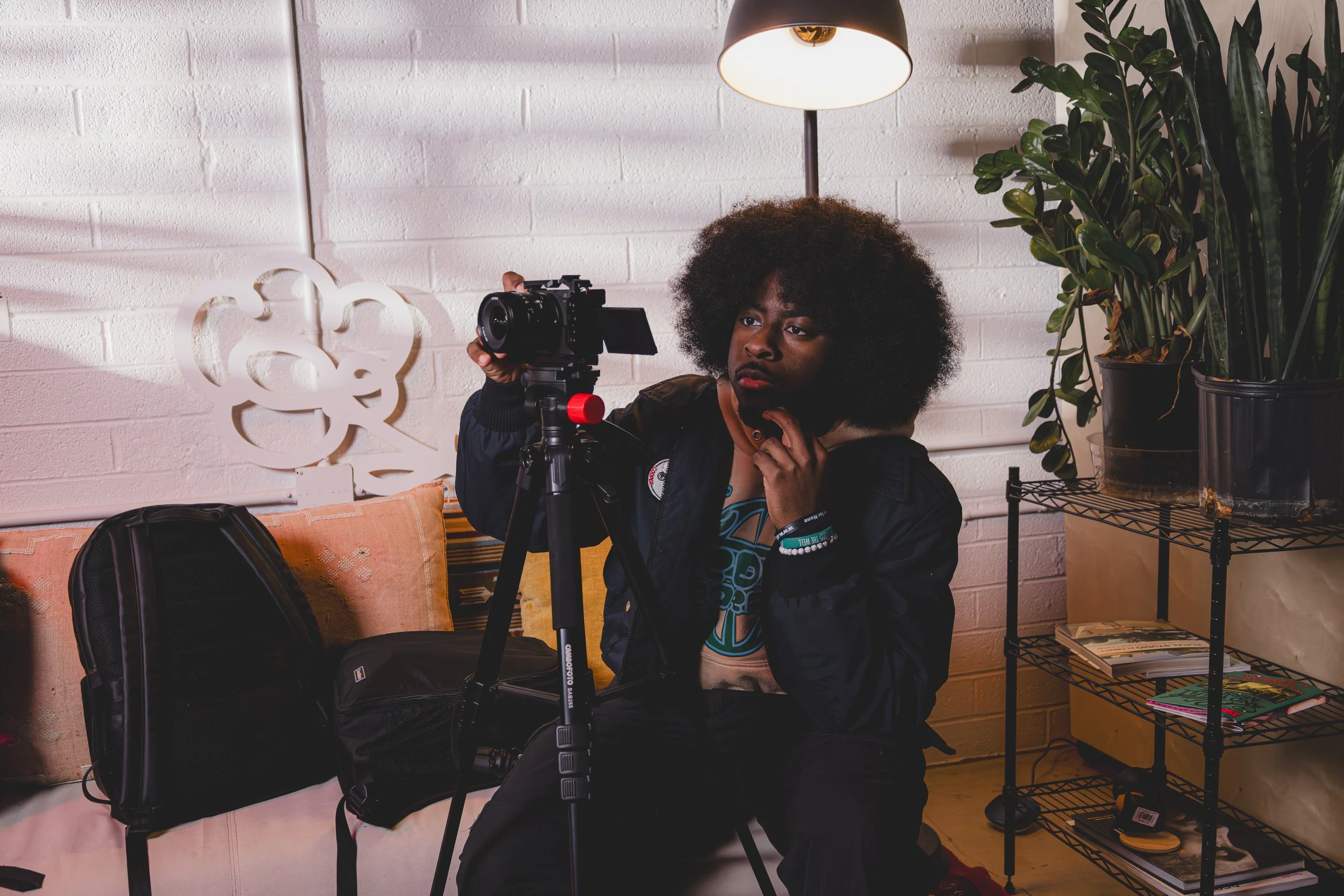 A woman with curly afro hair using a camera mounted on a tripod indoors, in front of a white brick wall with decorative white art and potted plants.