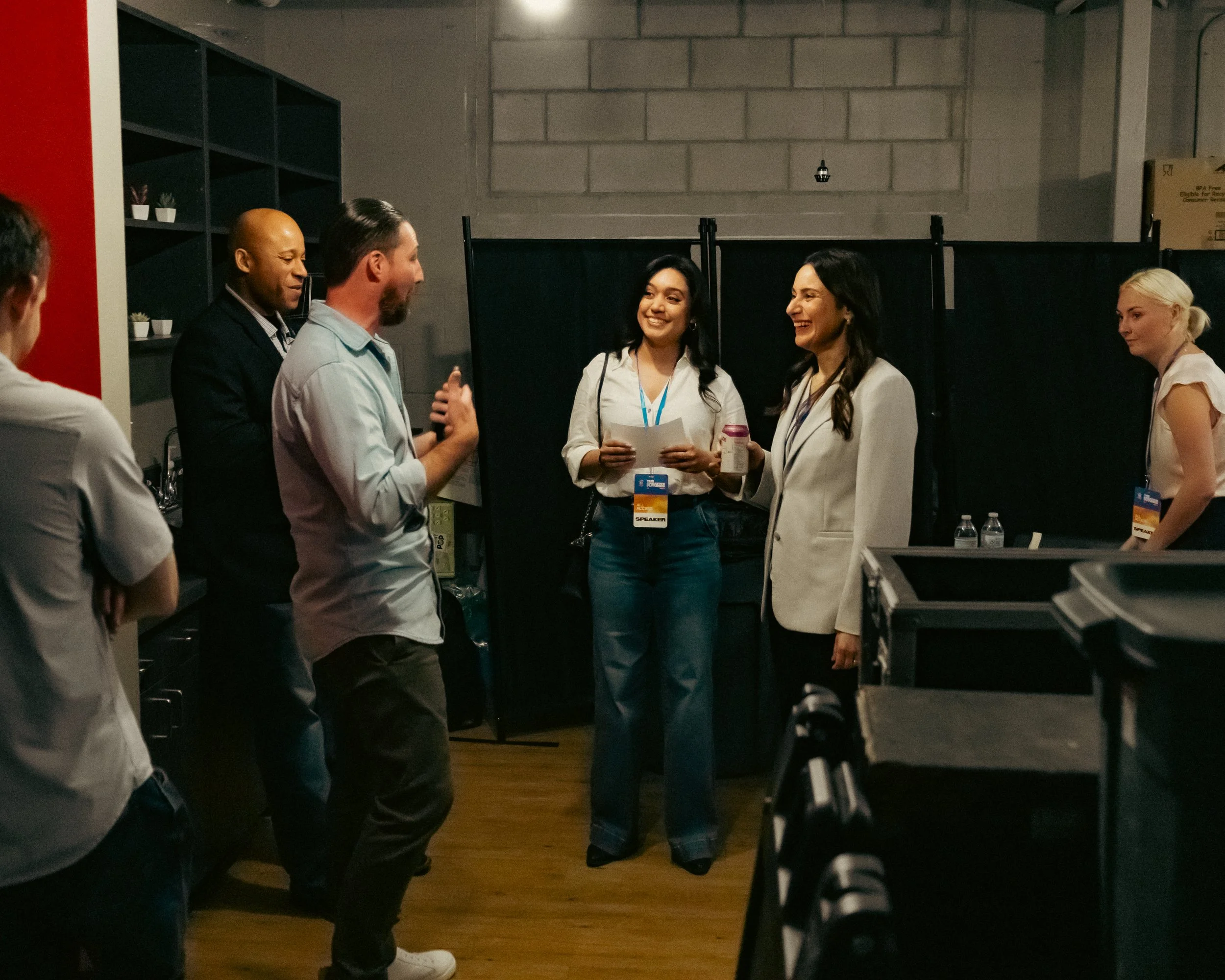 A group of six people standing and talking in a room with a black partition and shelves. The people are wearing business casual clothing, and some have conference name tags.
