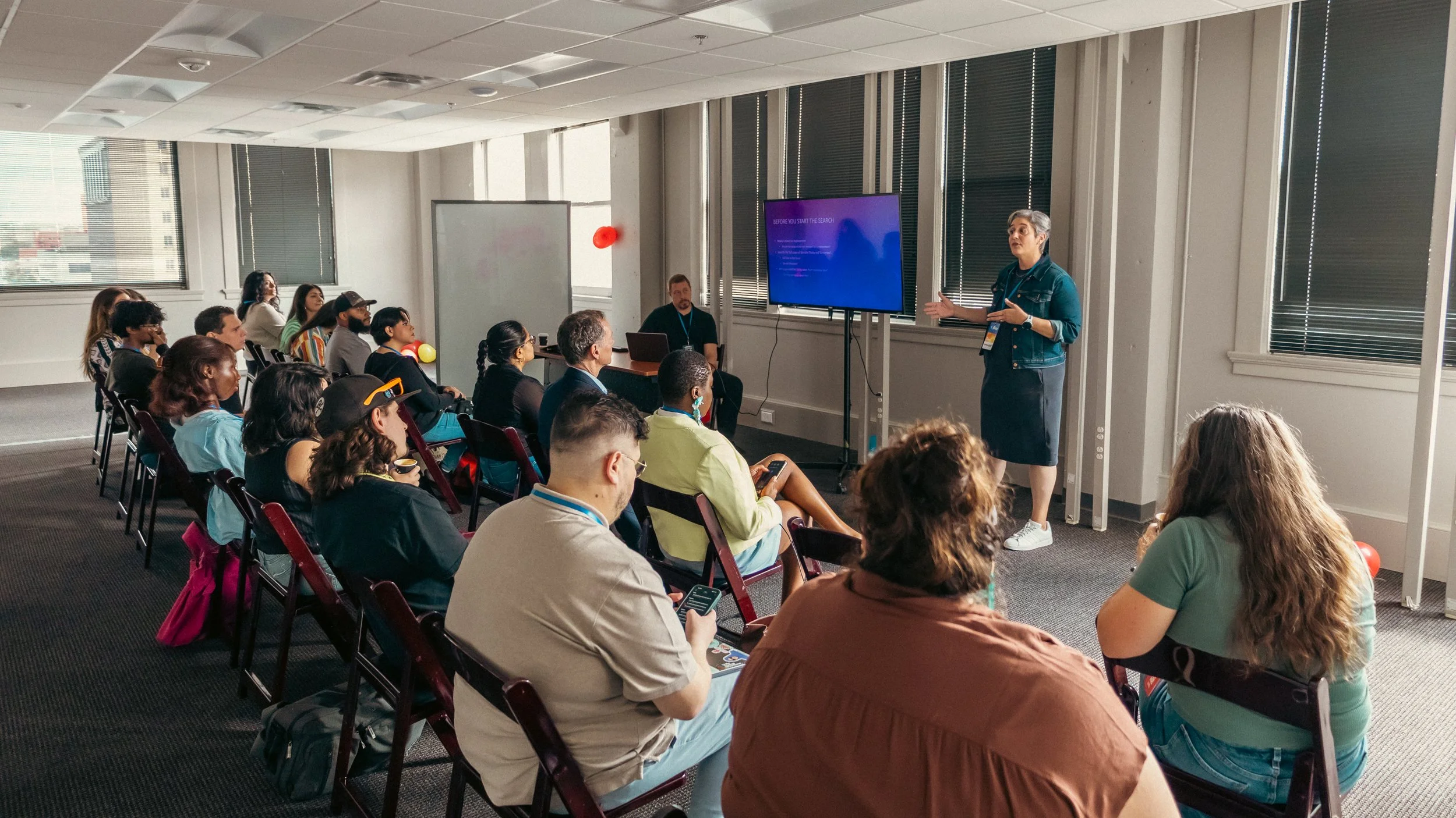 A woman giving a presentation to an audience seated in chairs in a conference room with large windows, blinds, and a screen displaying presentation slides.