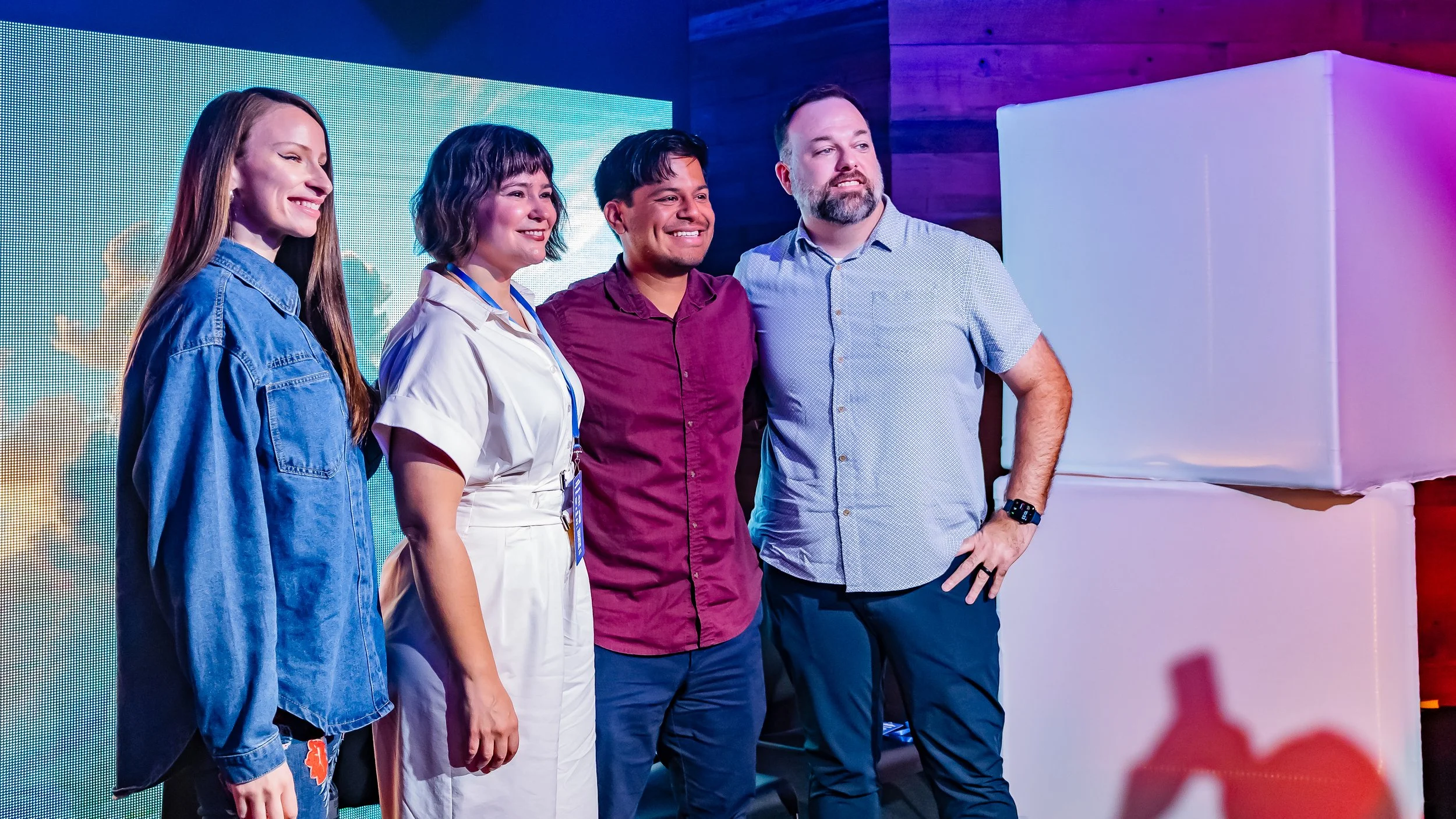 Four people standing on a stage in front of a bright digital screen, smiling and posing for a photo at an event.