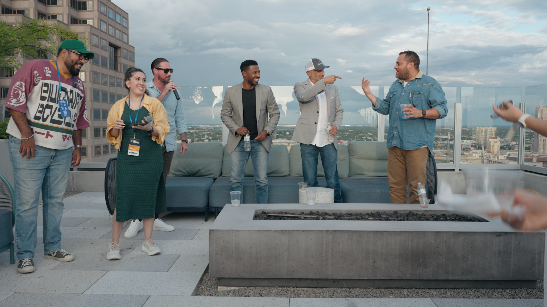 Group of seven diverse people on a rooftop, with some holding microphones, in casual and business attire, engaged in a conversation. The cityscape and partly cloudy sky are visible in the background.