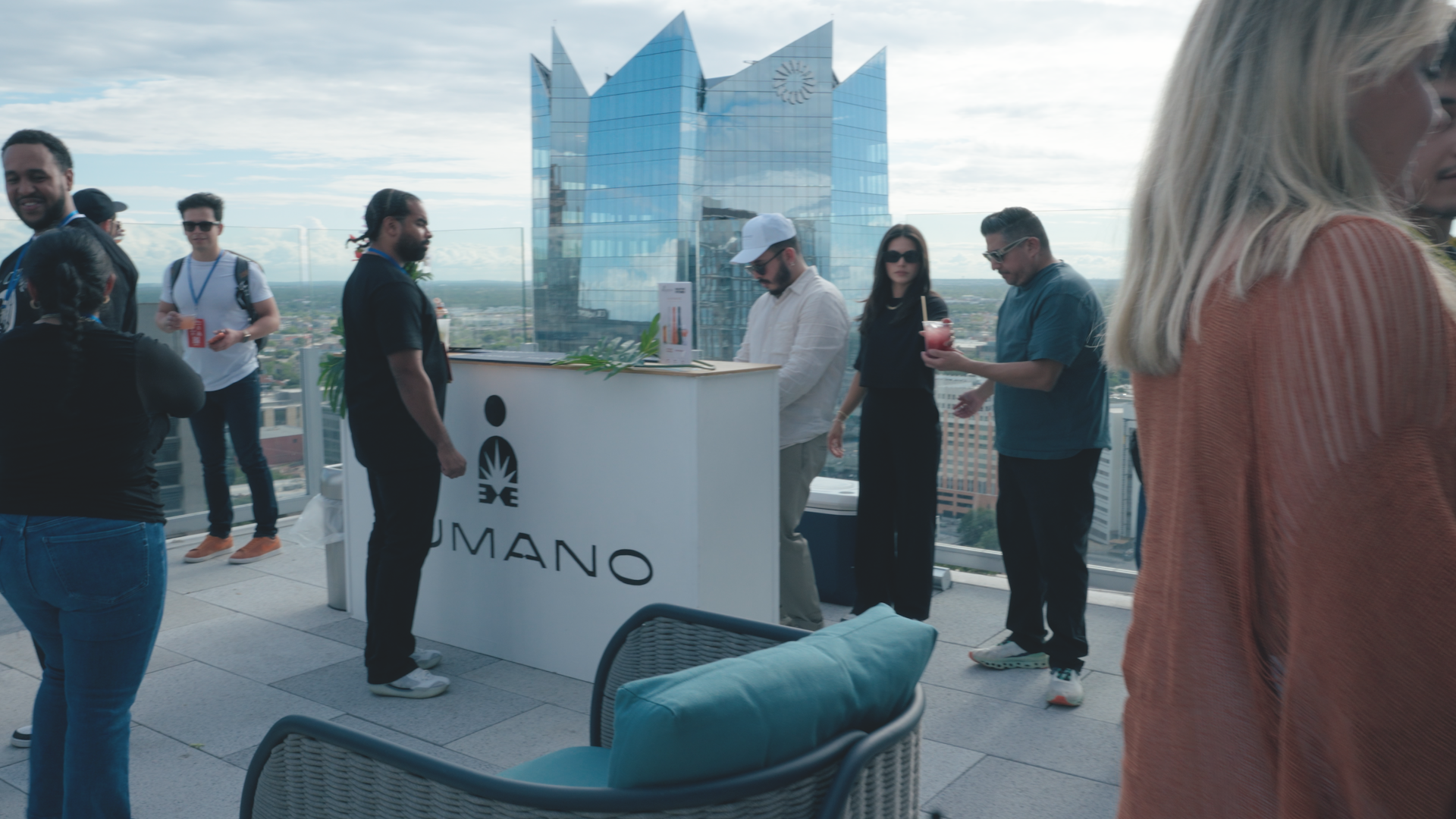 A group of people gathered on a rooftop terrace with city views, socializing and enjoying drinks, with a modern glass building in the background.
