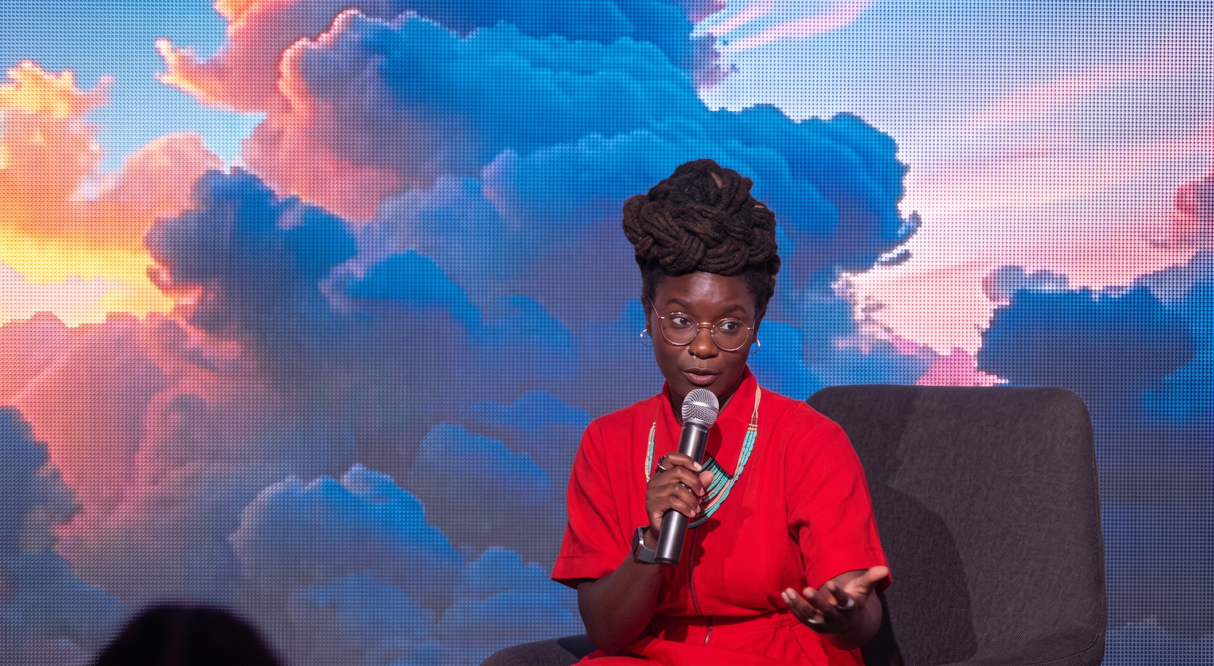 A woman with dark skin and large dreadlocks styled in an updo, wearing glasses and a red shirt, holding a microphone while speaking in front of a colorful cloud-like background on a screen.