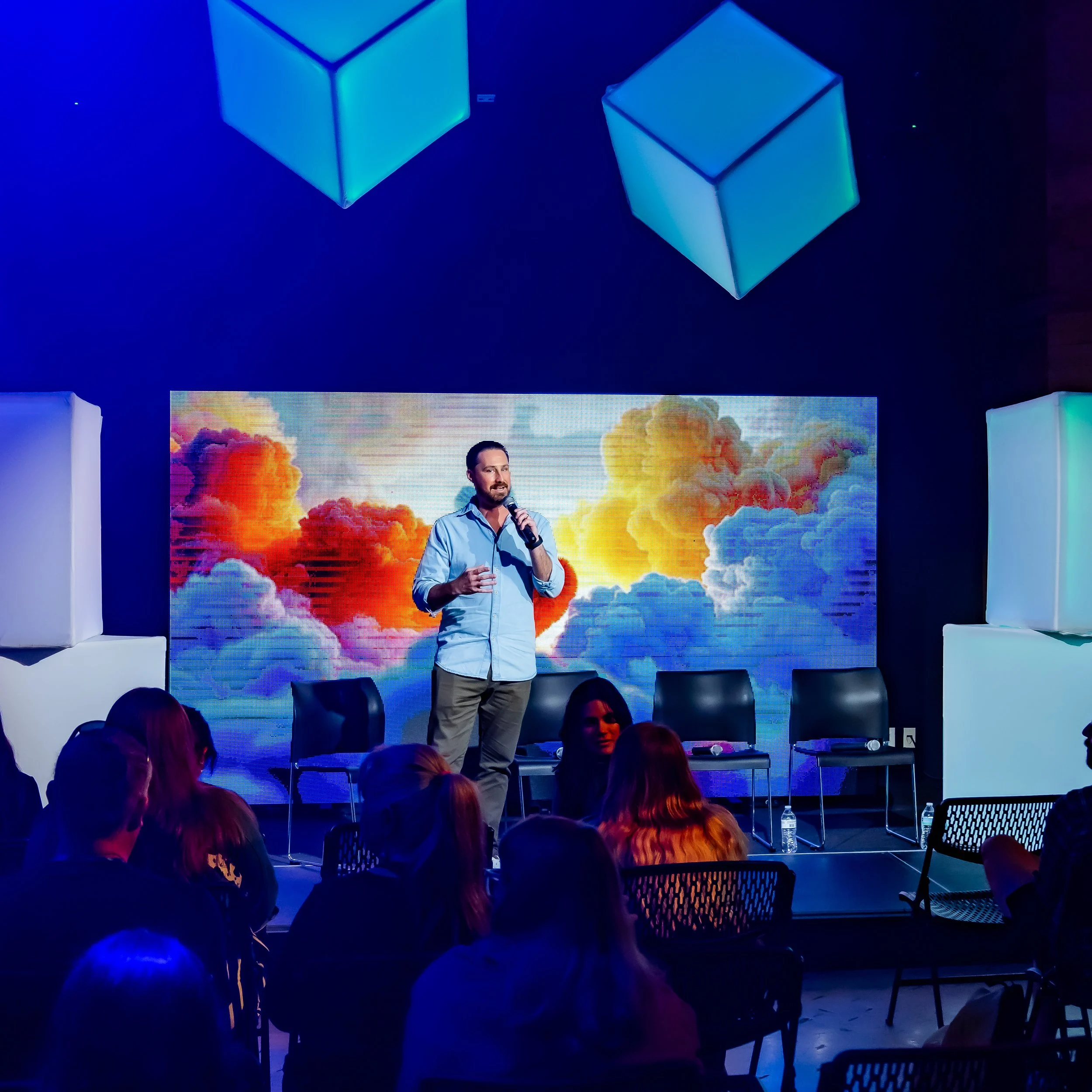 A man in a blue shirt holding a microphone stands on stage with a large colorful cloud background on a screen behind him. The stage is decorated with white block-like structures and there is an audience seated facing the stage.