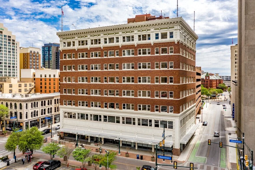 A large multi-story brick building on a city street with trees, cars, and other buildings nearby, under a partly cloudy sky.