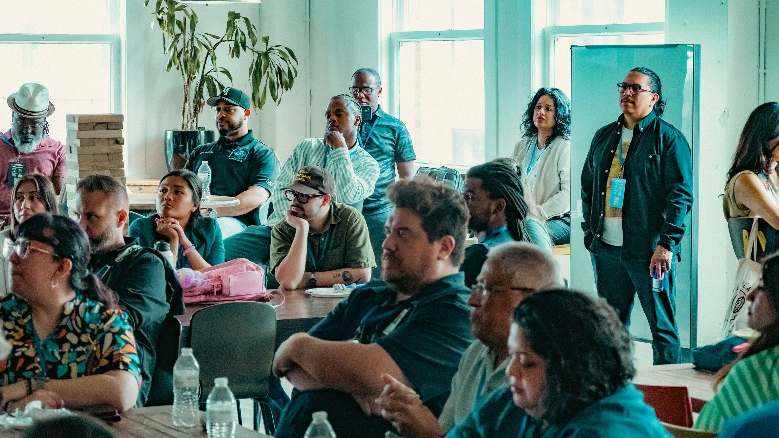 A diverse group of people attending a conference or seminar in a brightly lit room with large windows. Some are seated, while others are standing and listening attentively.