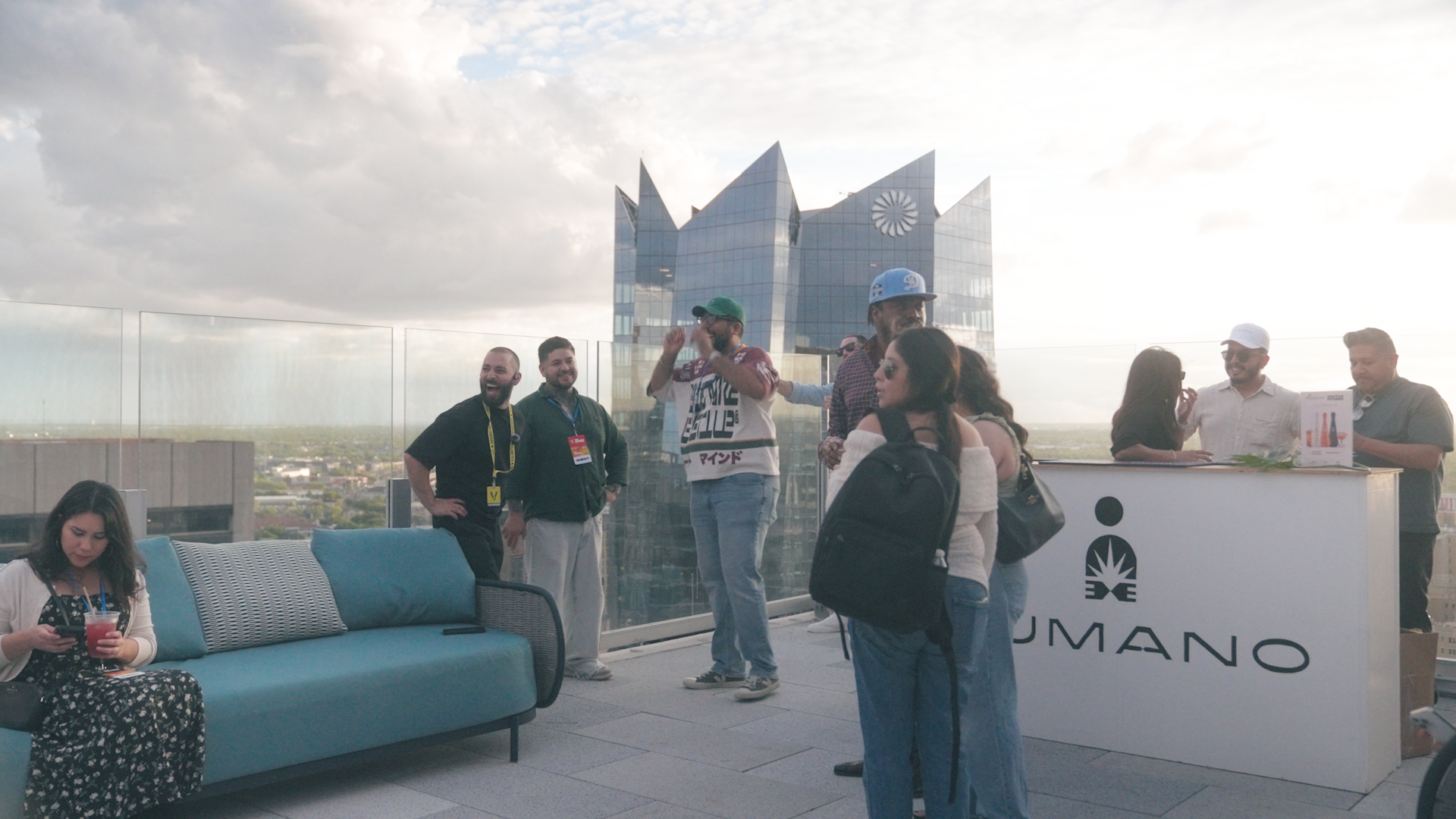 People socializing on a rooftop terrace with a city view and modern building in the background; a woman in a floral dress using her phone with a drink in her hand, and a group around a bar area.
