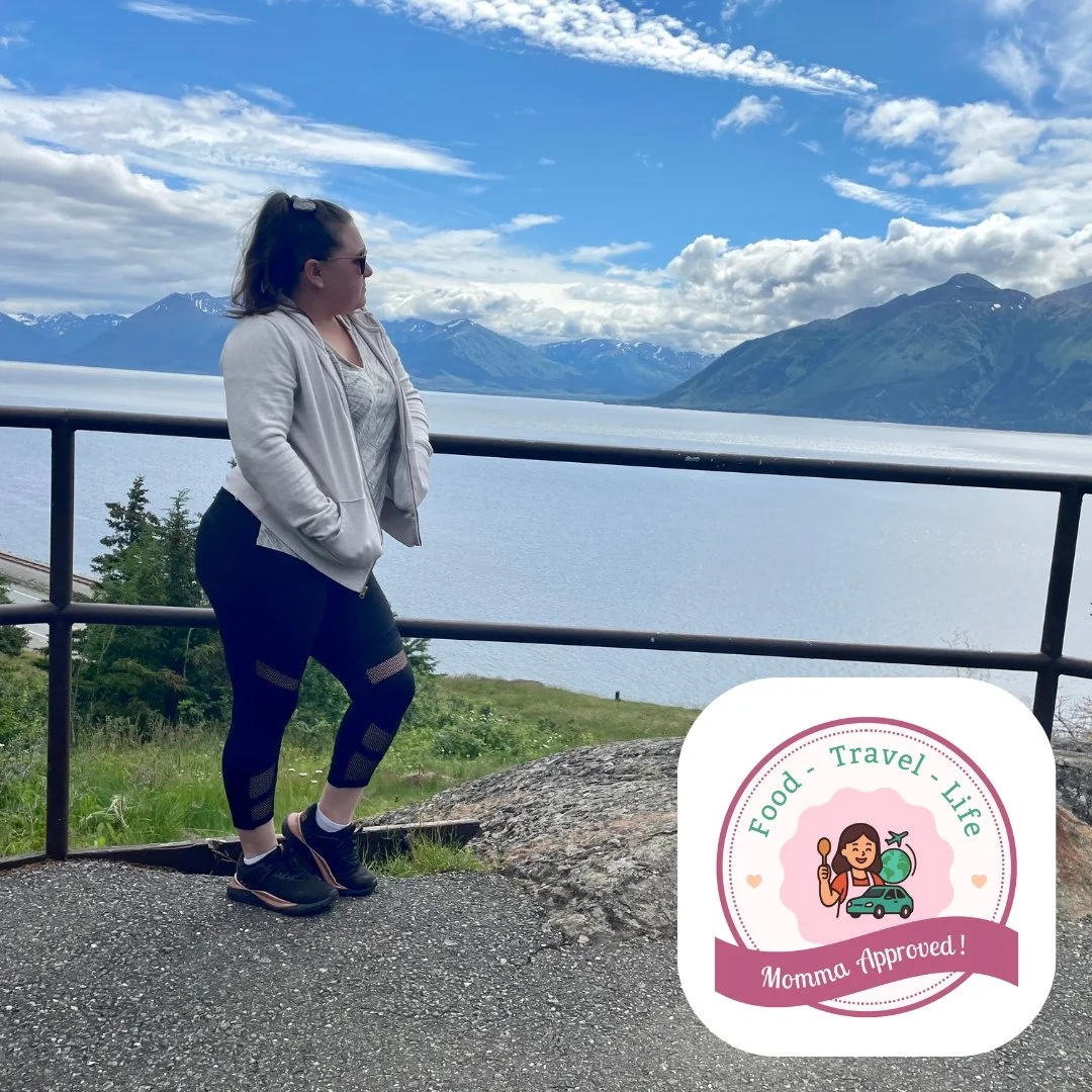 Woman in athletic clothes and sunglasses leaning on a railing at a scenic lake with Alaskan mountains and a cloudy sky in the background.
