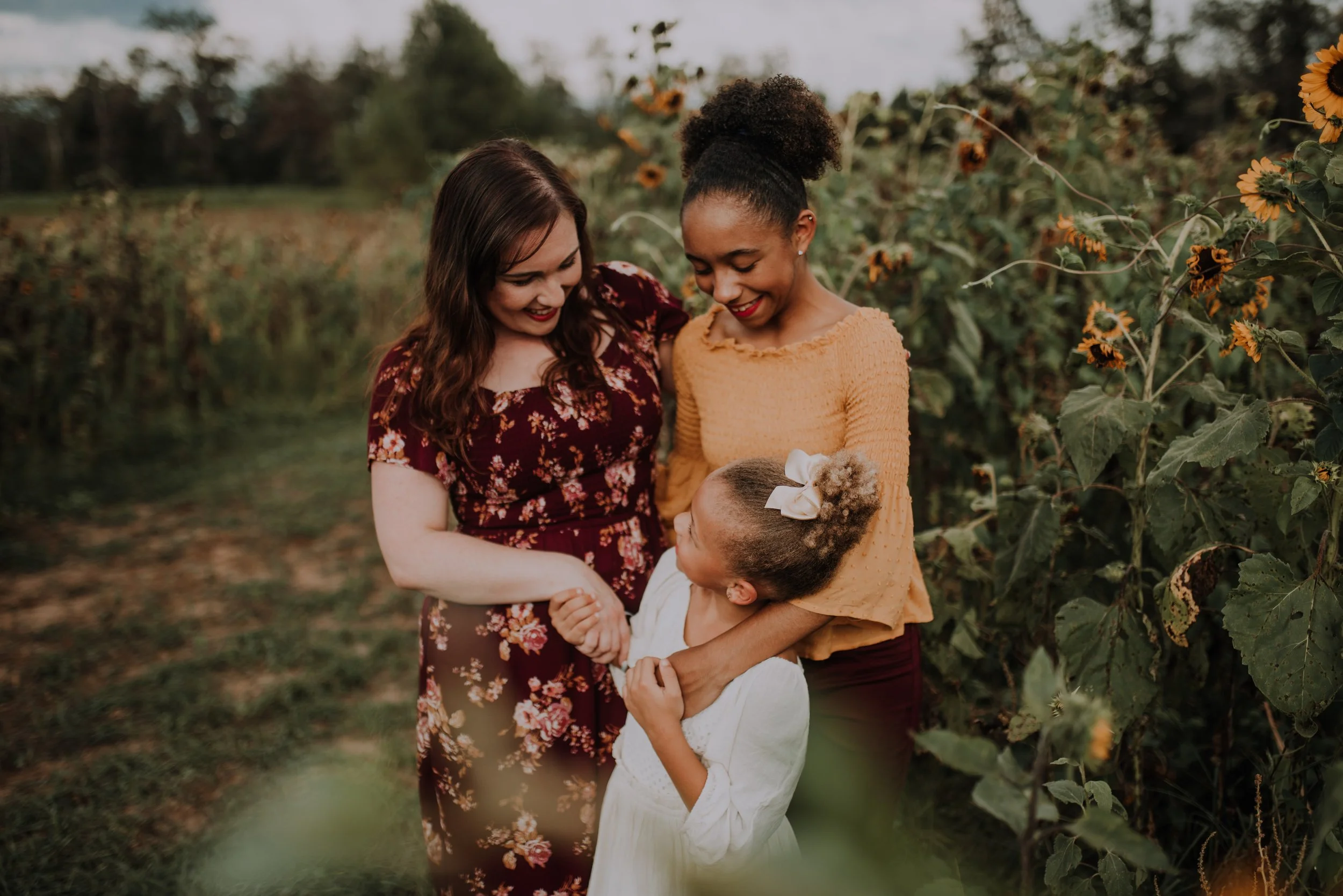 Mom and daughters hugging in a sunflower field with greenery and sunflowers in the background.