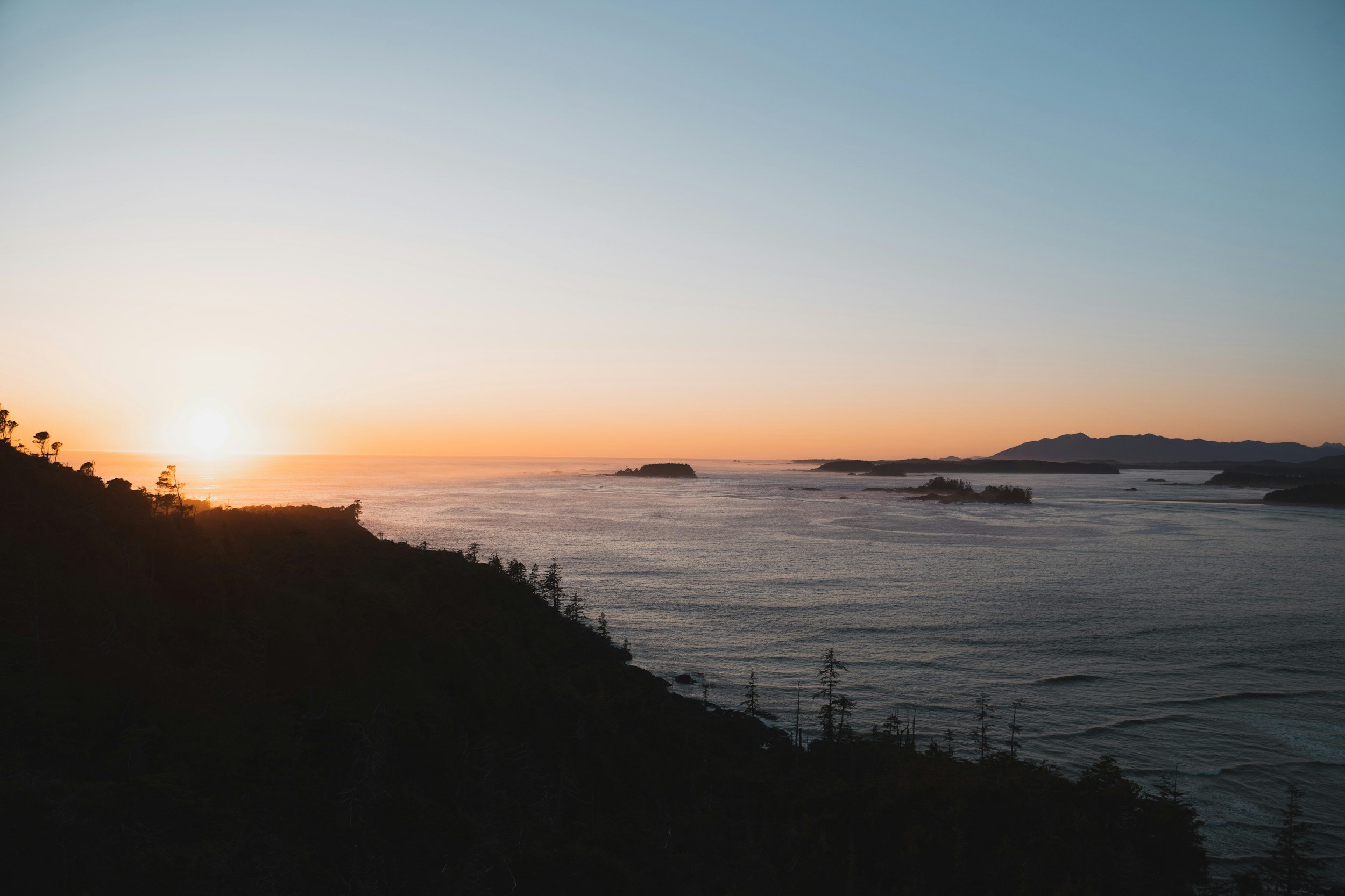 Sunset over the ocean with a silhouette of trees and distant mountains.