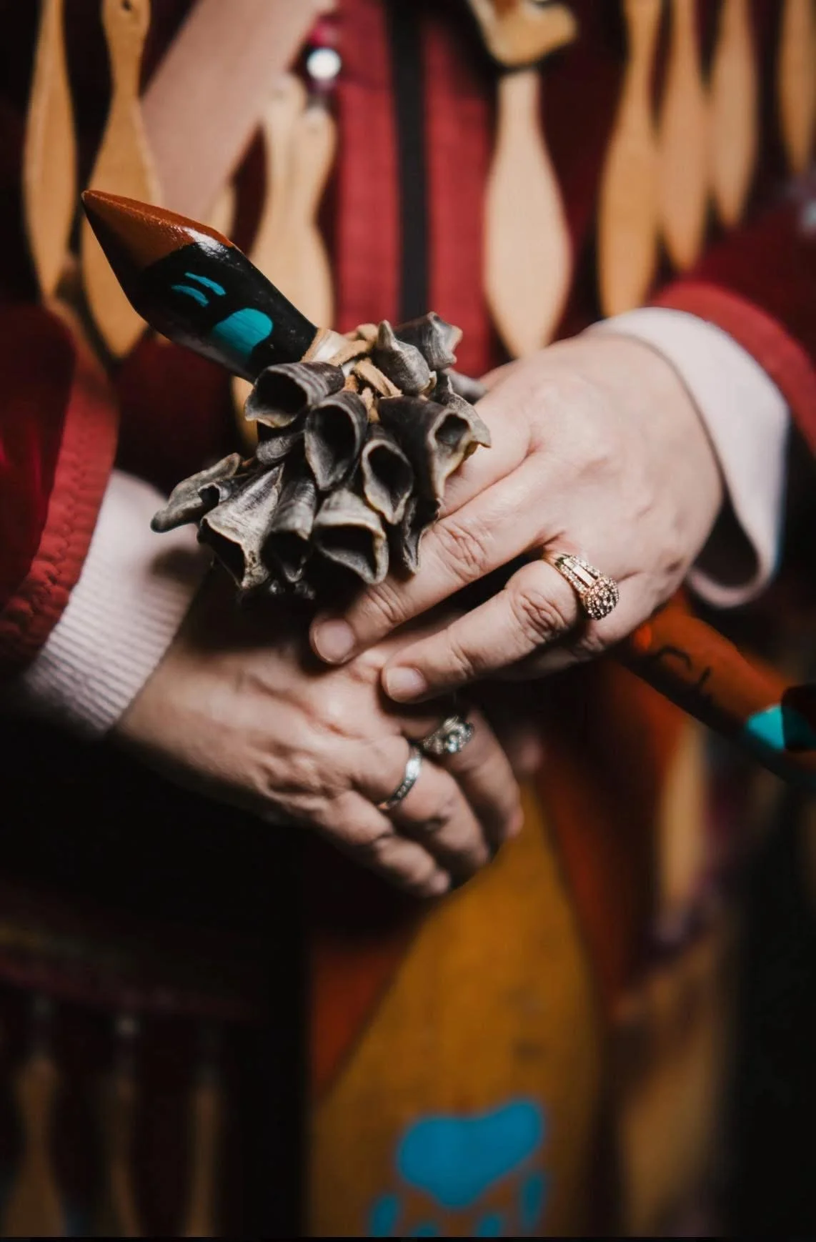 Close-up of a woman holding a bouquet of sagebrush and a large pencil with painted face features, wearing multiple rings on her fingers, against a blurred background with wooden decor and red outfit.