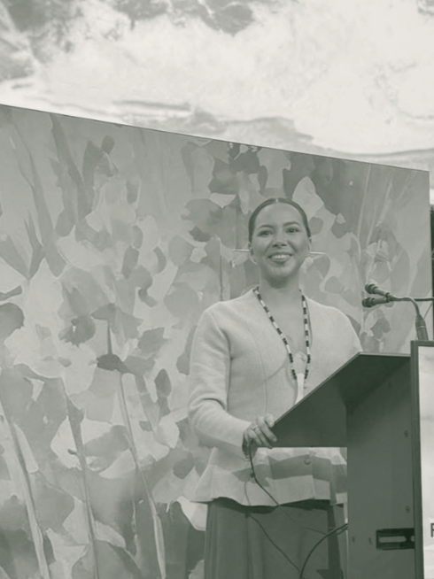 A woman speaking at a podium during an outdoor event with a patterned backdrop and cloudy sky.