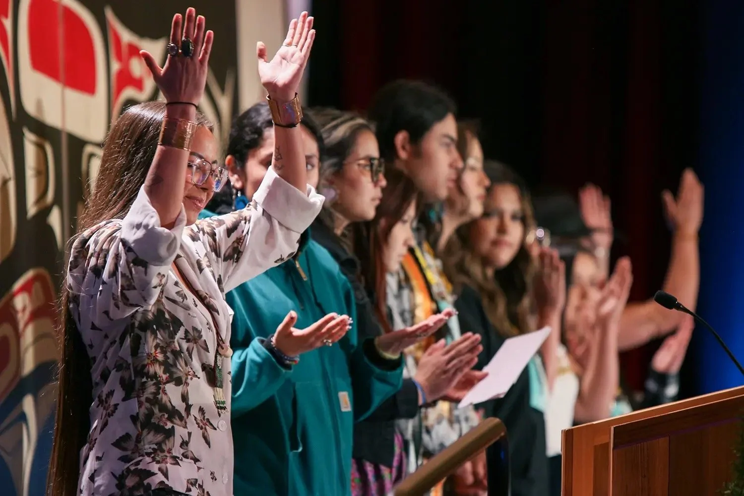 A diverse group of people standing at a podium, raising their right hands, participating in an oath or pledge ceremony, with some holding papers.